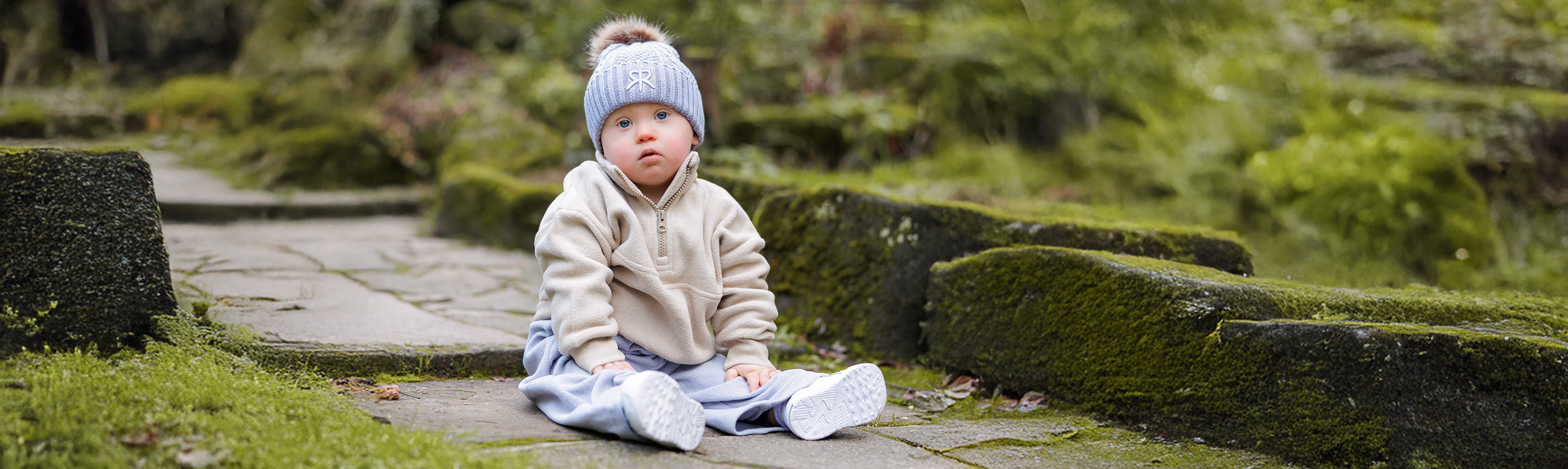 Toddler sitting on a stone path in a garden in winter with knit beanie and fleece, cosy tracksuit