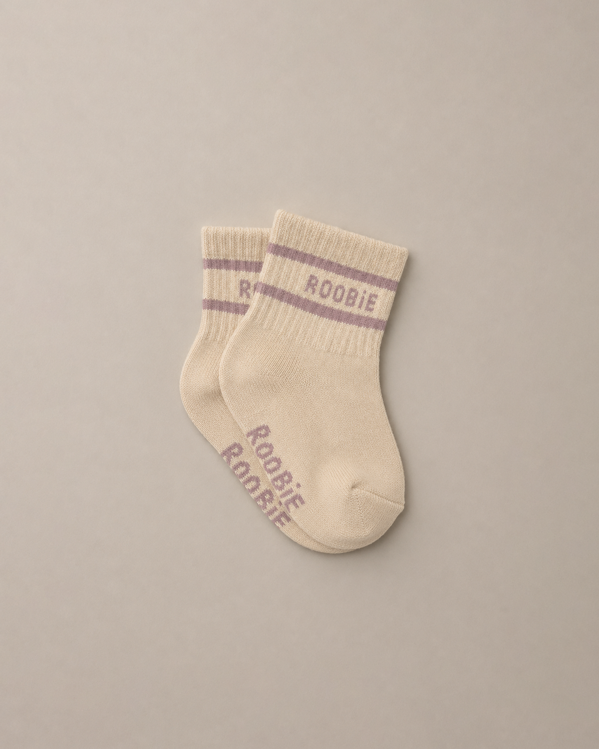 A pair of cream-colored baby socks with pink-purple stripes and 'ROOBIE' printed on the cuff and sole, displayed on a neutral beige background.