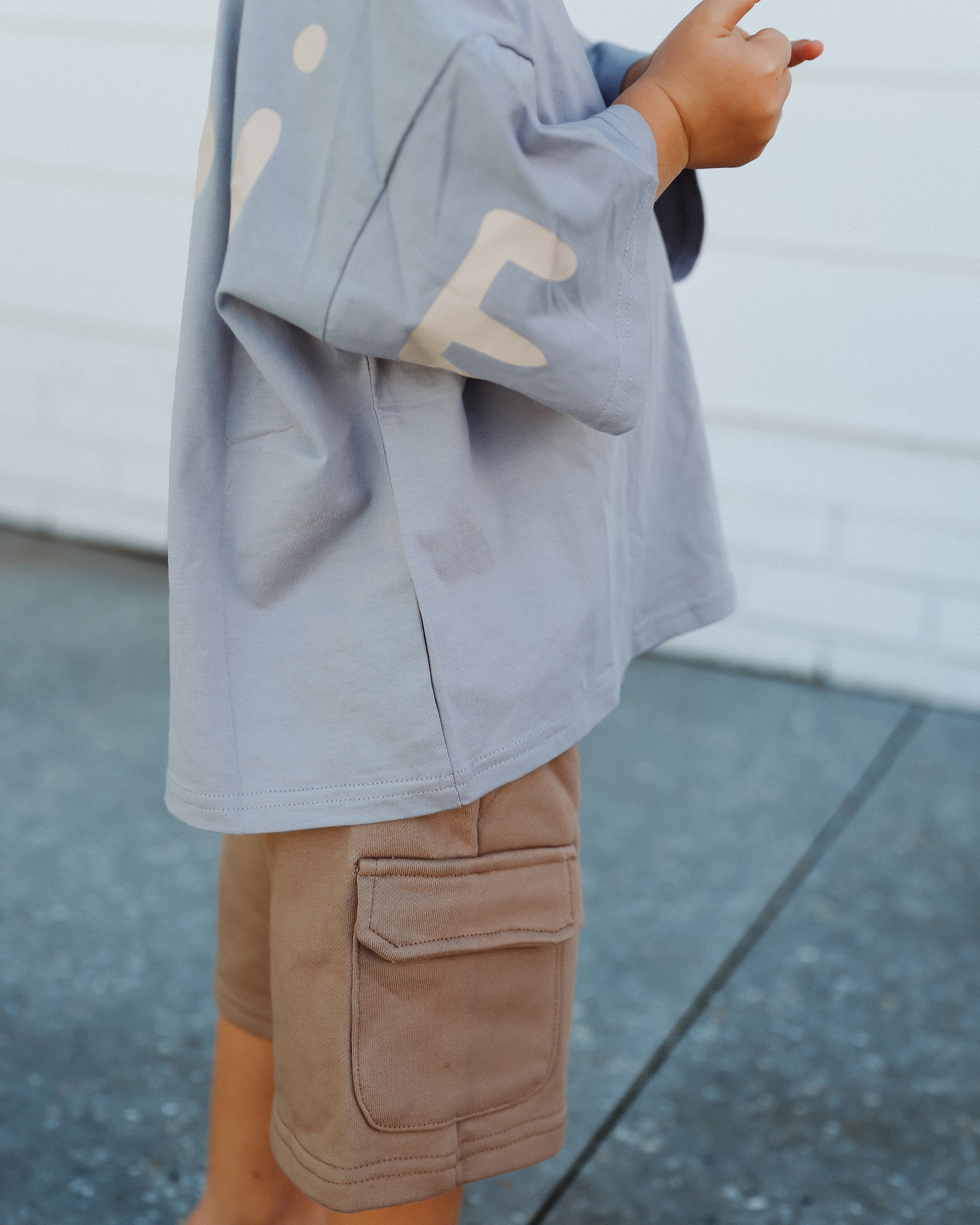 A young child mixing and matching a light blue Oversized T-shirt and brown Cargo Shorts, standing outdoors in front of a neutral white building.