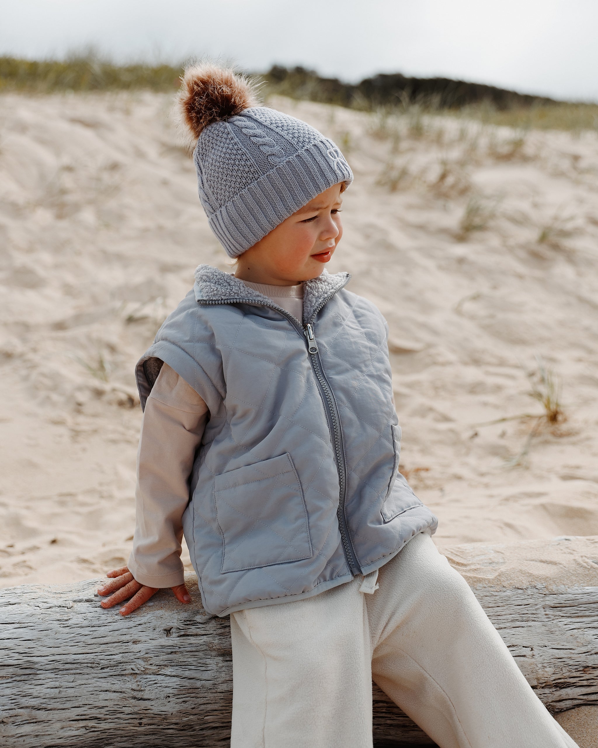 A toddler sitting on a log on a sandy beach dune, wearing a light blue-gray quilted gilet 4-in-1 reversible coat, a matching knit bobble beanie, and cream fleece wide leg joggers.