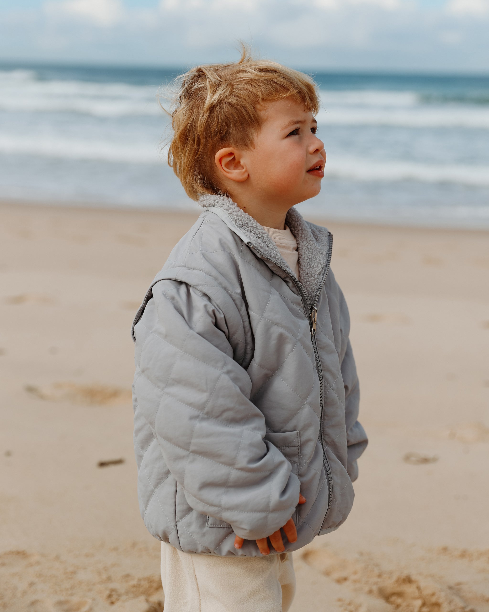 A young child wearing a light gray-blue quilted 4-in-1 reversible teddy coat and cream wide leg pants, standing on a sandy beach with the ocean and cloudy sky in the background.