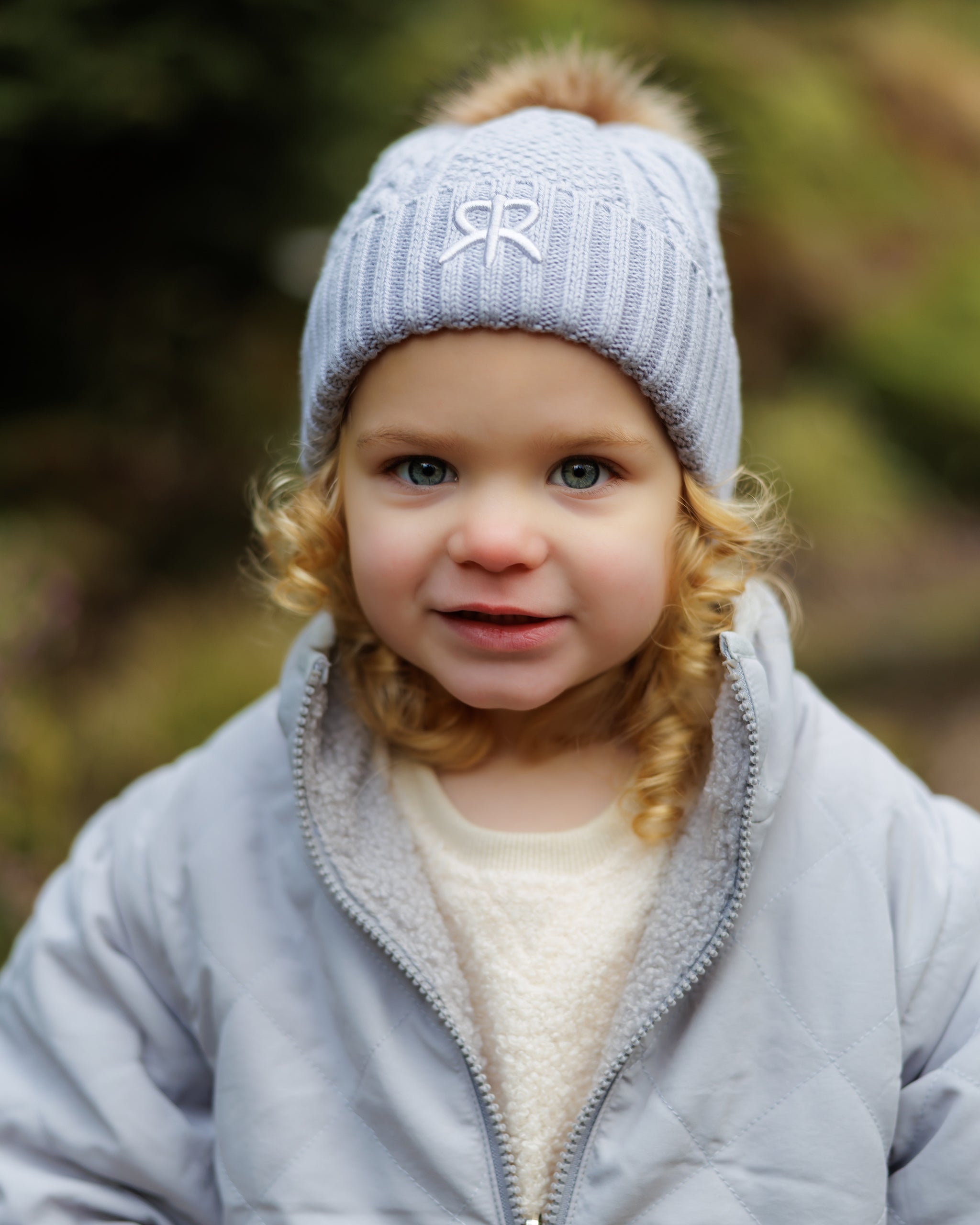 Child wearing a blue knit bobble beanie hat with a logo and blue quilted 4-in-1 jacket outdoors