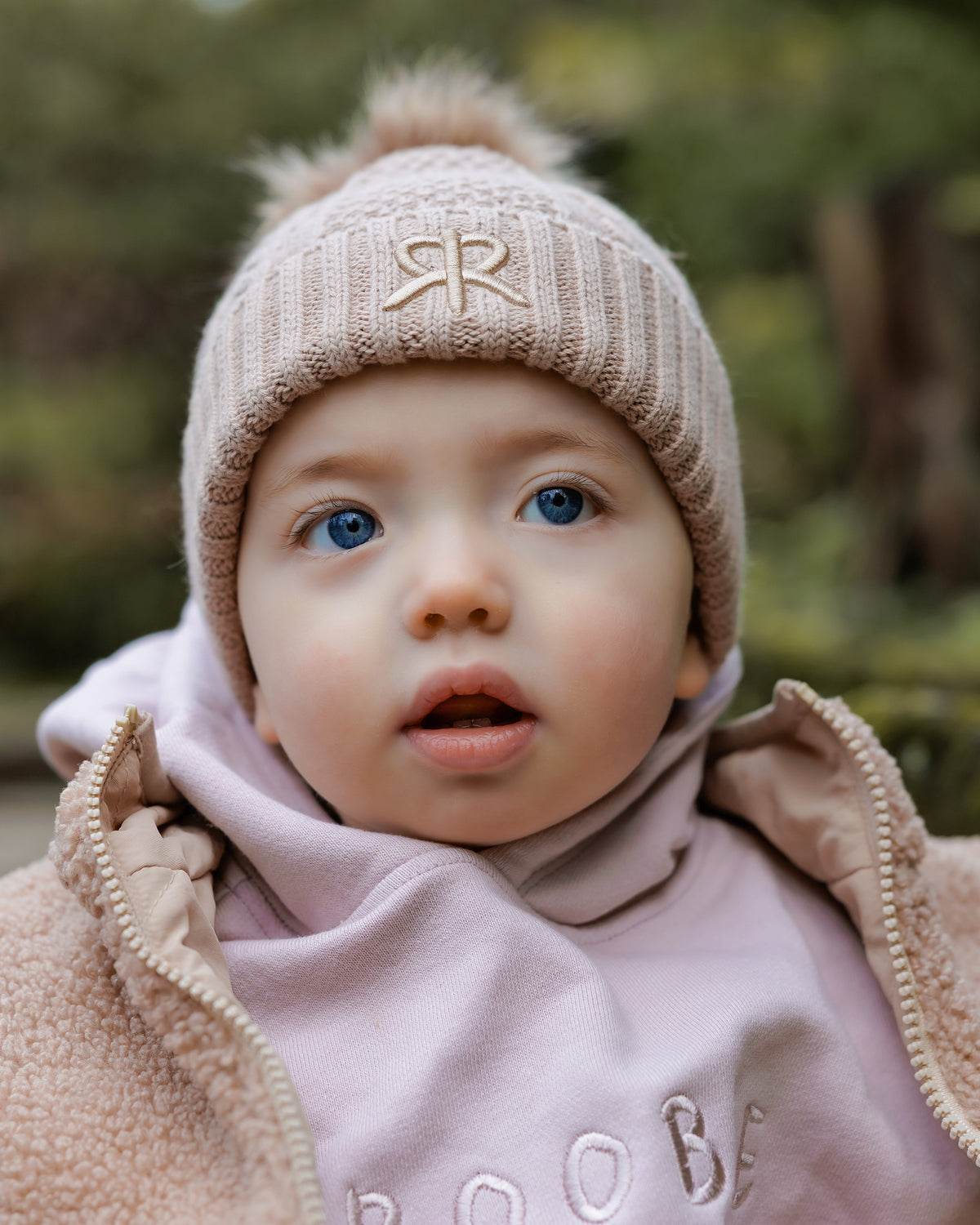 Child wearing a brown knit bobble beanie hat with a logo and pink oversized hoodie and brown teddy coat outdoors
