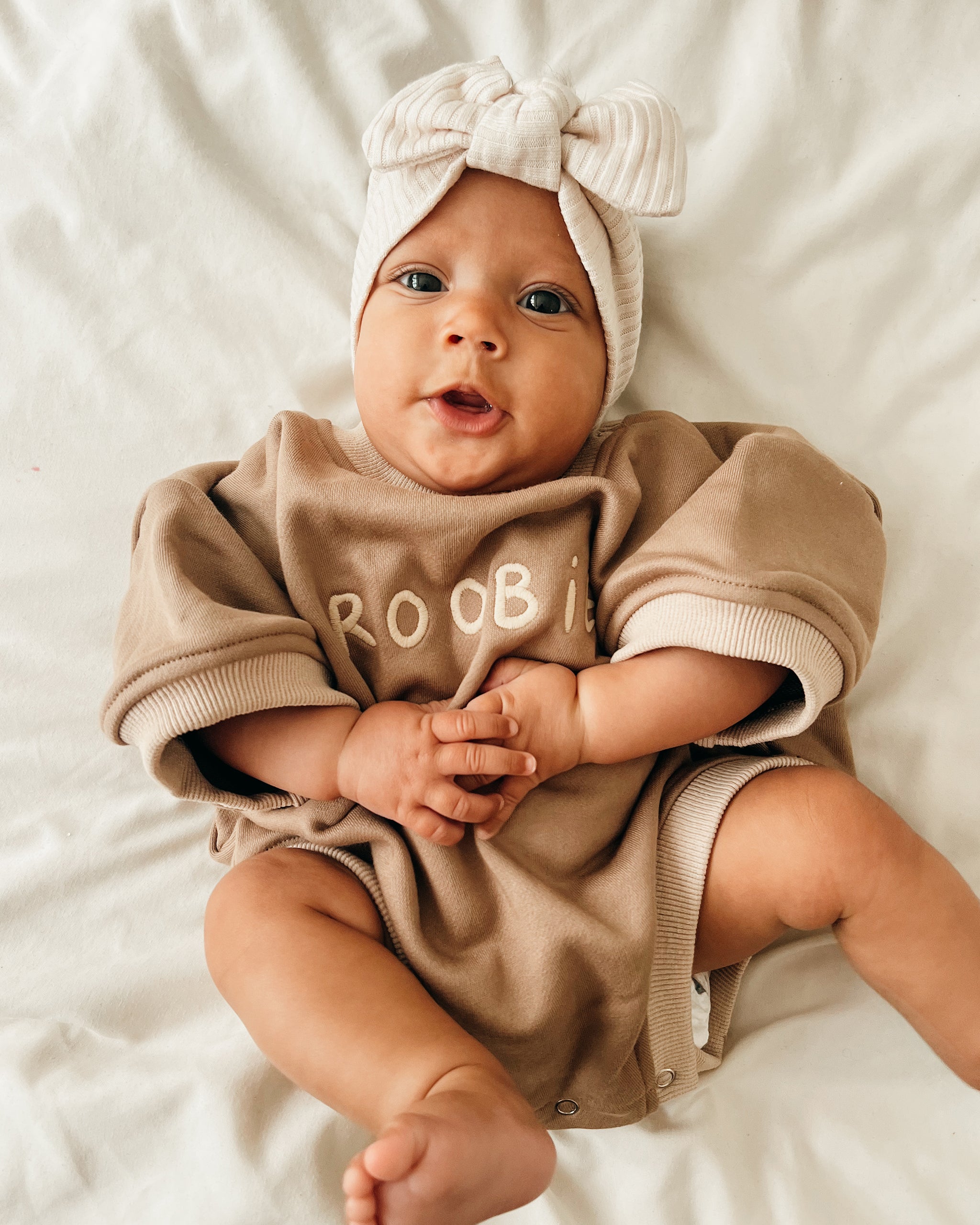 A baby lying on white bedding, wearing a beige and brown neutral oversized romper with 'ROOBiE' printed on the chest and a matching beige ribbed bow headband.