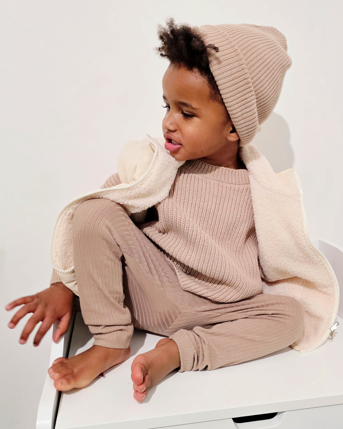 Child wearing a brown chunky knit sweater, with matching brown ribbed cotton beanie, leggings and cream quilted gilet sitting on a white surface