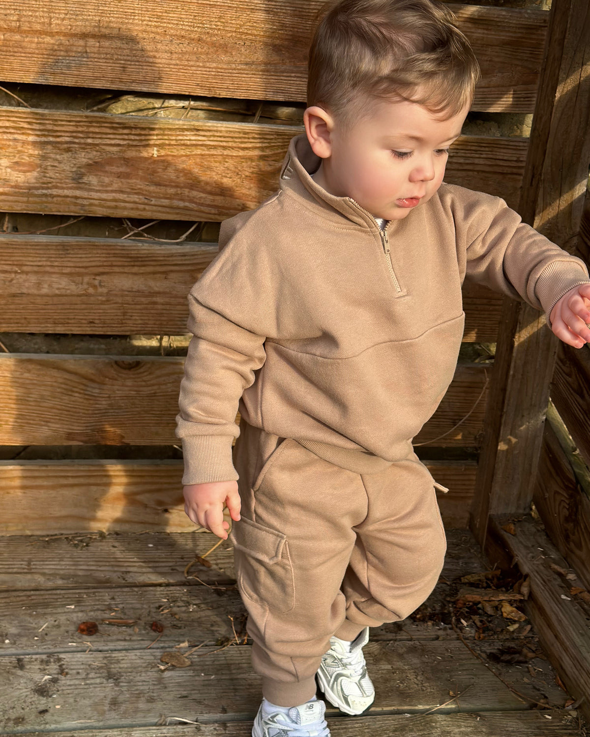 Child in a brown tracksuit with quarter zip and matching cargo pants standing against a wooden wall