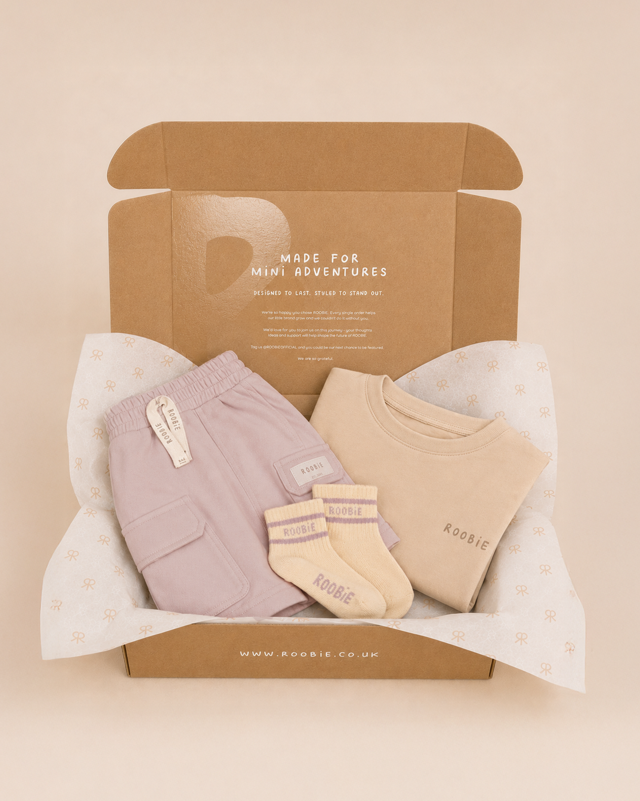 A box showing ROOBiE pink cargo shorts, cream and pink striped crew socks and cream oversized t-shirt on a beige studio backdrop with the words 'made for mini adventures' on the box