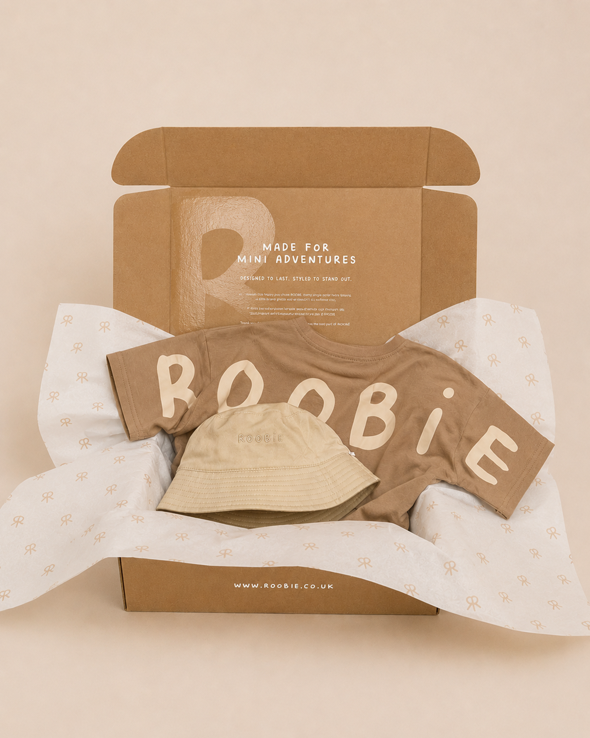 A box showing ROOBiE oversized brown t-shirt and reversible cream and brown bucket hat on a beige studio backdrop with the words 'made for mini adventures' on the box