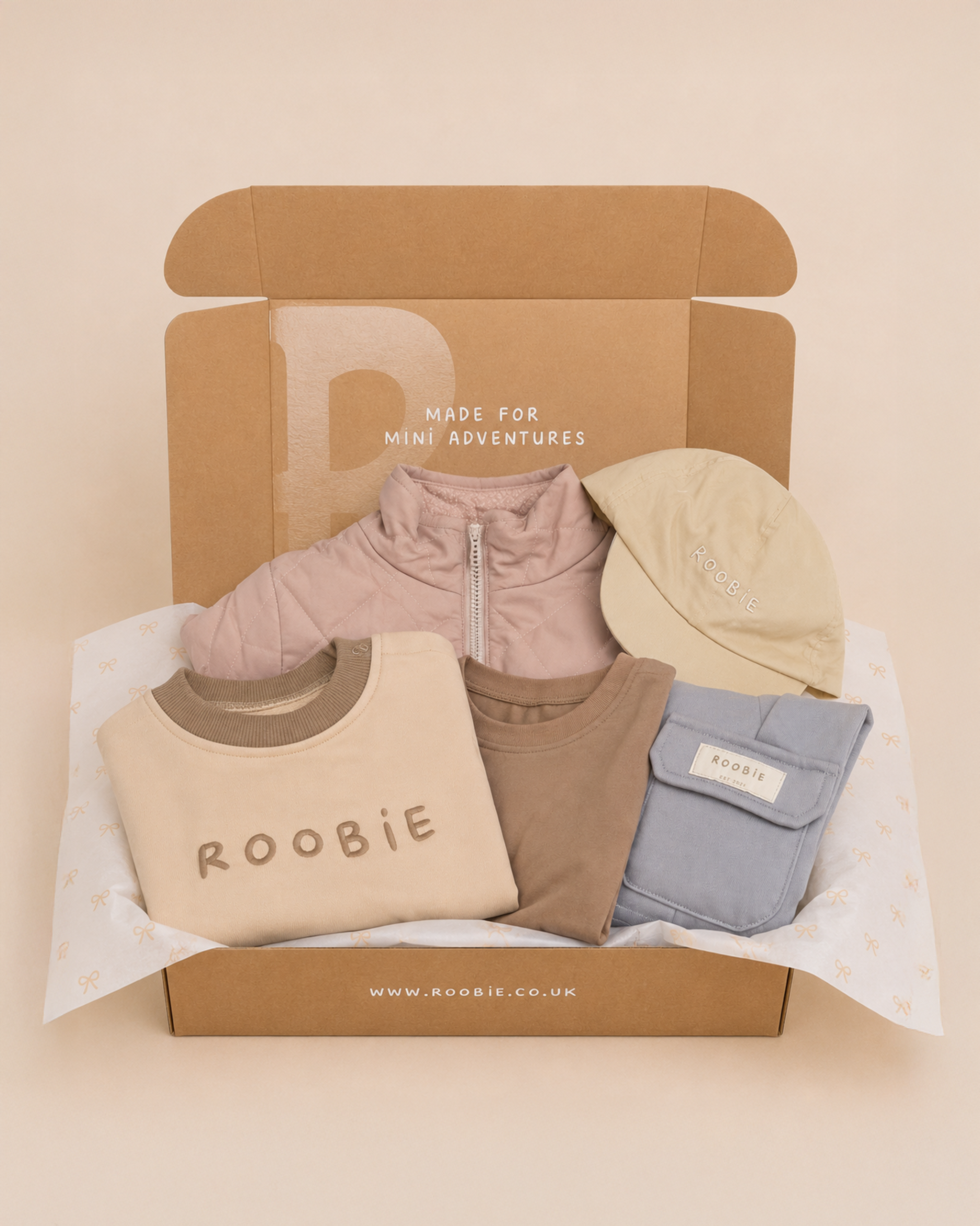 A box showing ROOBiE oversized romper in cream, pink 4-in-1 reversible coat, brown jumper, blue cargos, reversible cap in cream on a beige studio backdrop with the words 'made for mini adventures' on the box