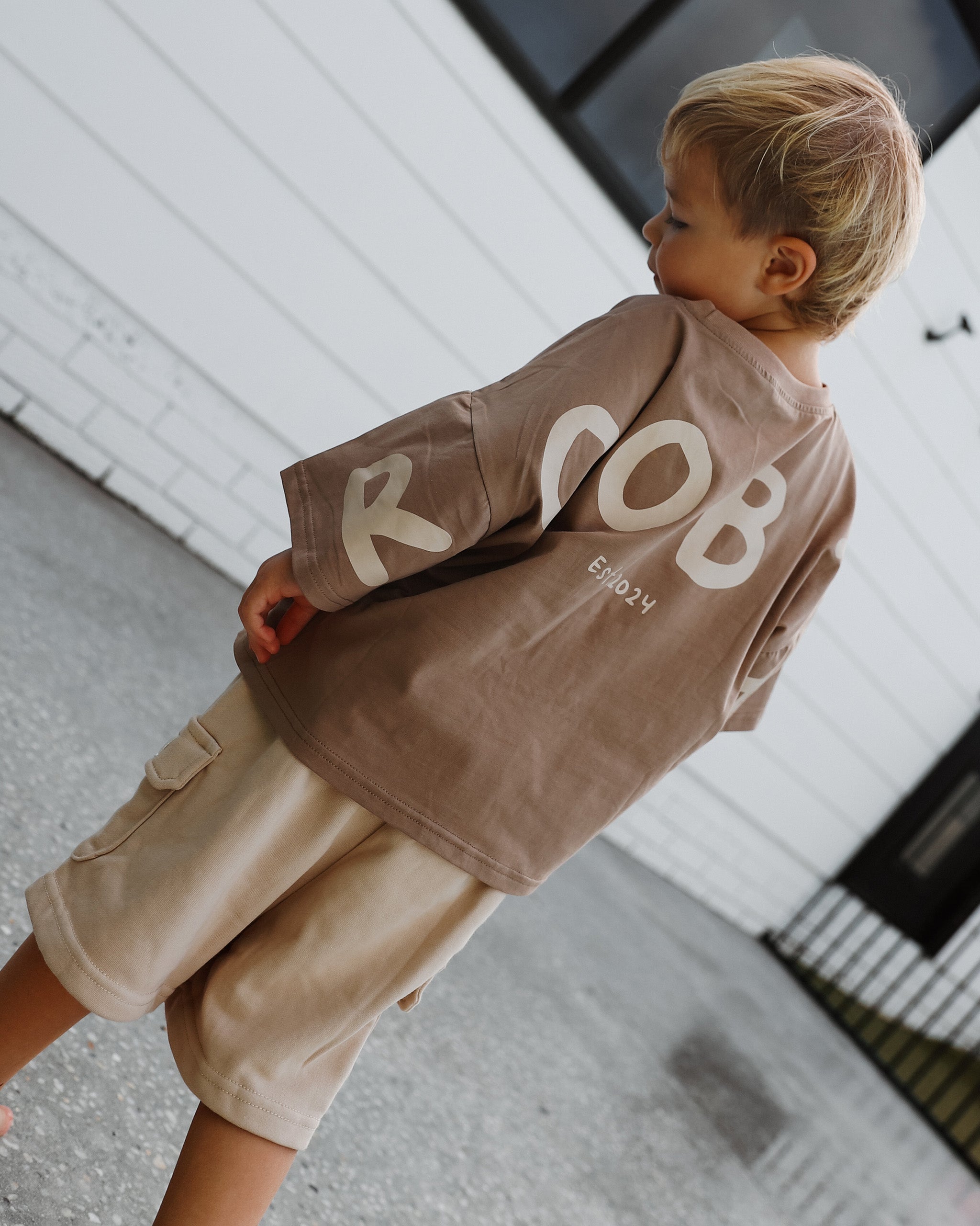 A young child wearing a brownish-beige oversized T-shirt with large cream letters on the back and beige Cargo Shorts, photographed outdoors against a white wall and gray pavement.