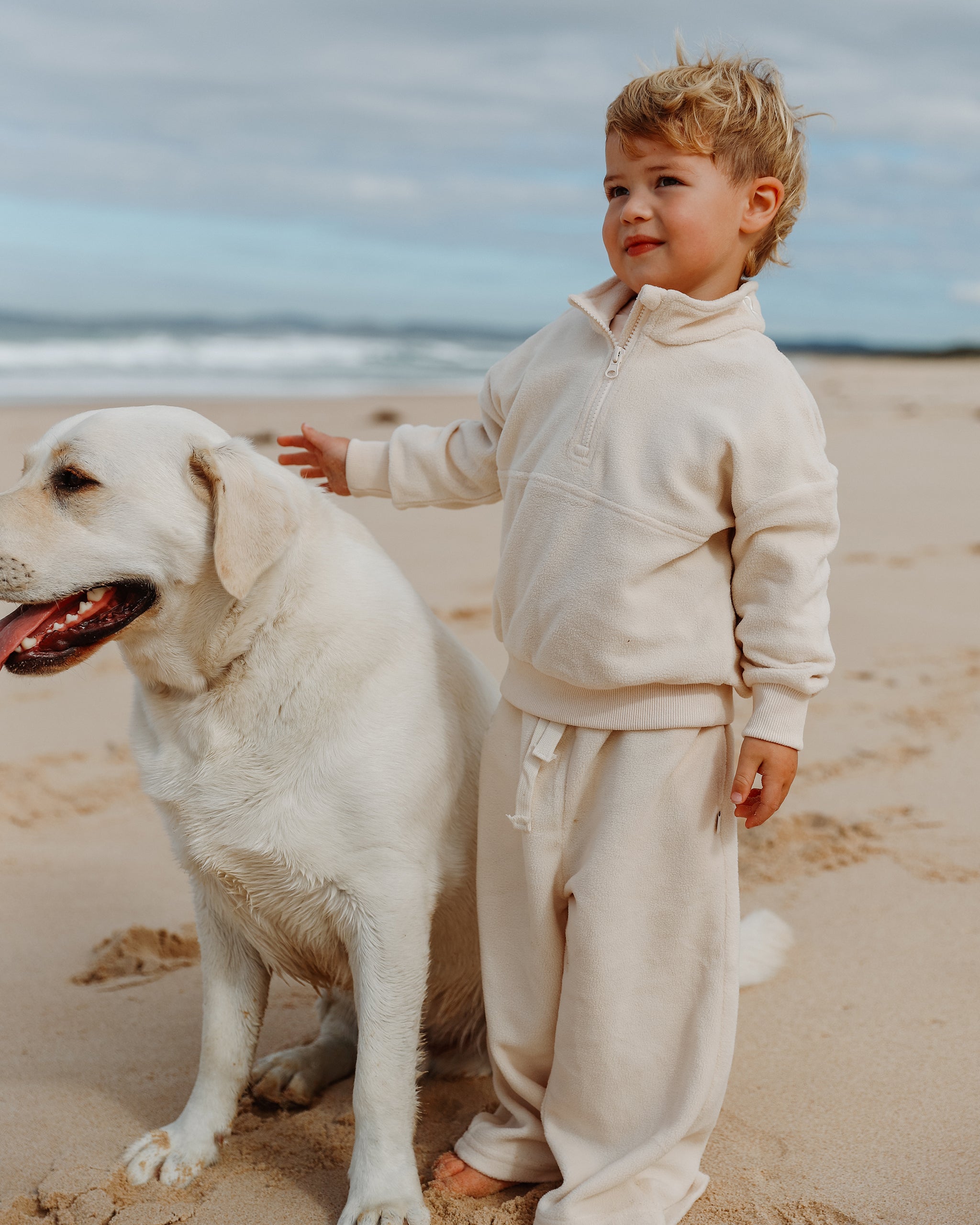 A toddler wearing a cream-colored matching zip-up sweatshirt and sweatpants stands on a sandy beach next to a large white dog, with the ocean and cloudy sky in the background.
