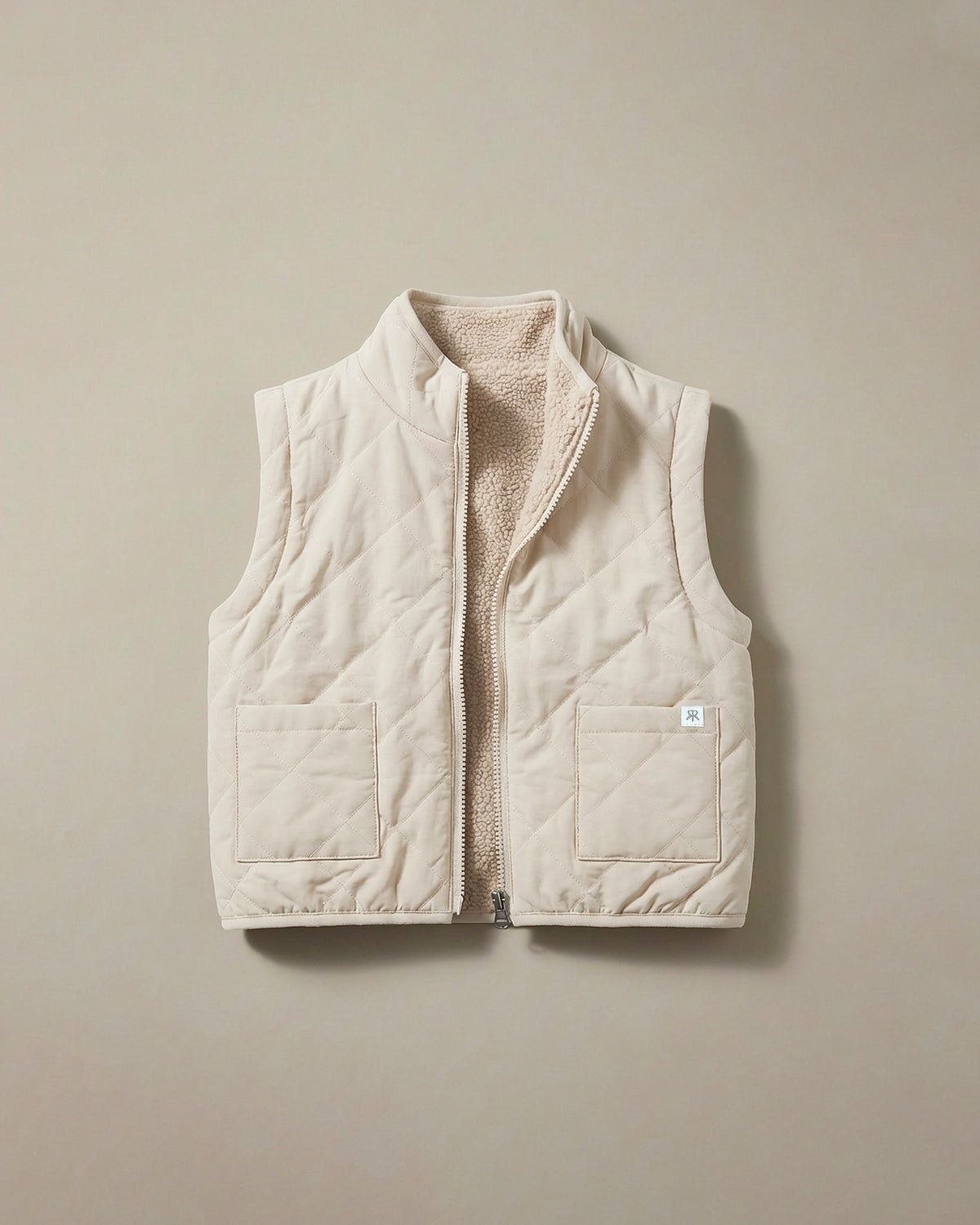 A light beige quilted puffer vest with a soft teddy fleece lining, shown flat against a neutral beige background.