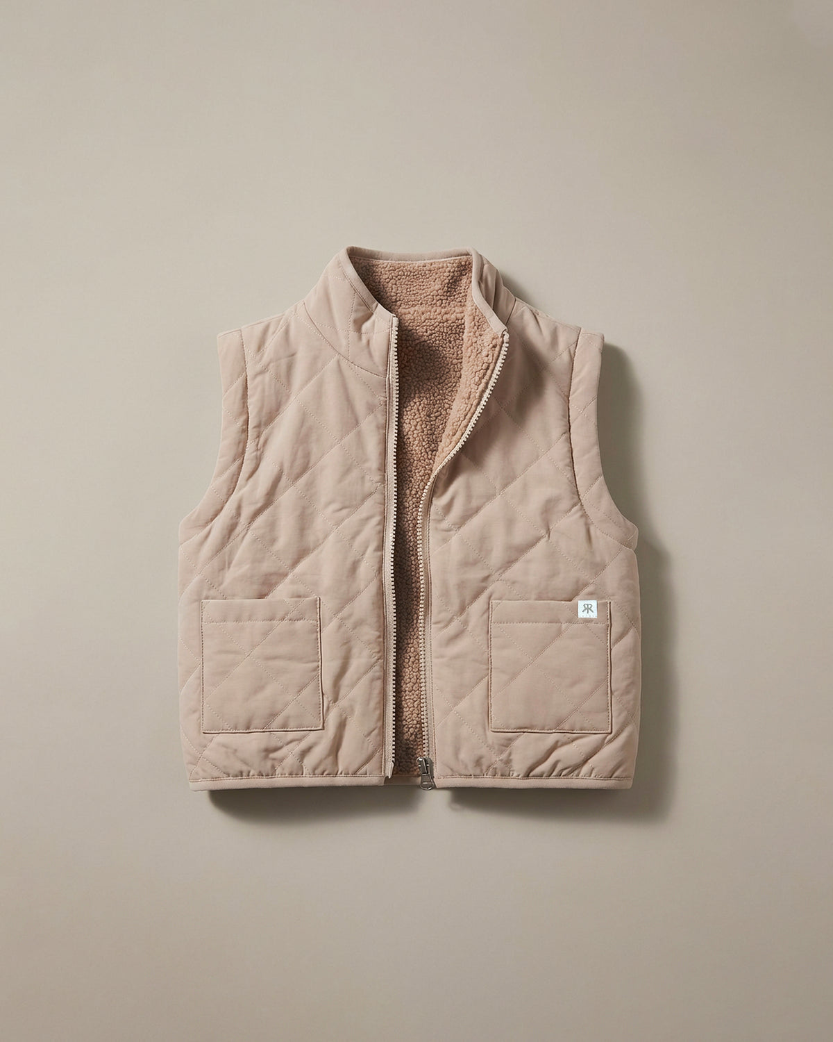 A light beige quilted sleeveless gilet with a soft tan fleece lining, shown flat against a neutral beige background.