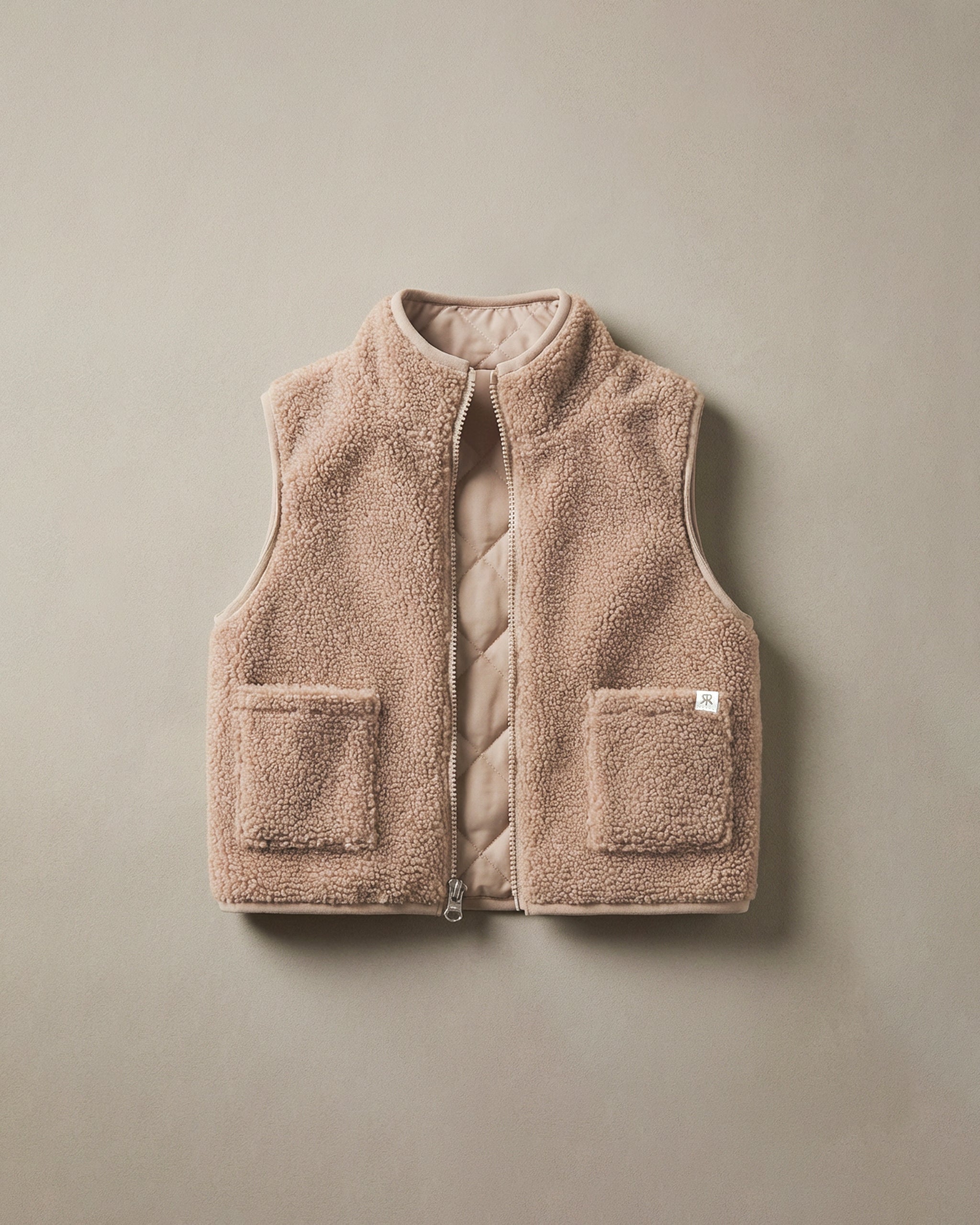 A sleeveless teddy fleece gilet in a warm taupe-beige color, featuring a quilted interior lining, a front zipper, and two front patch pockets, shown flat against a neutral beige background.