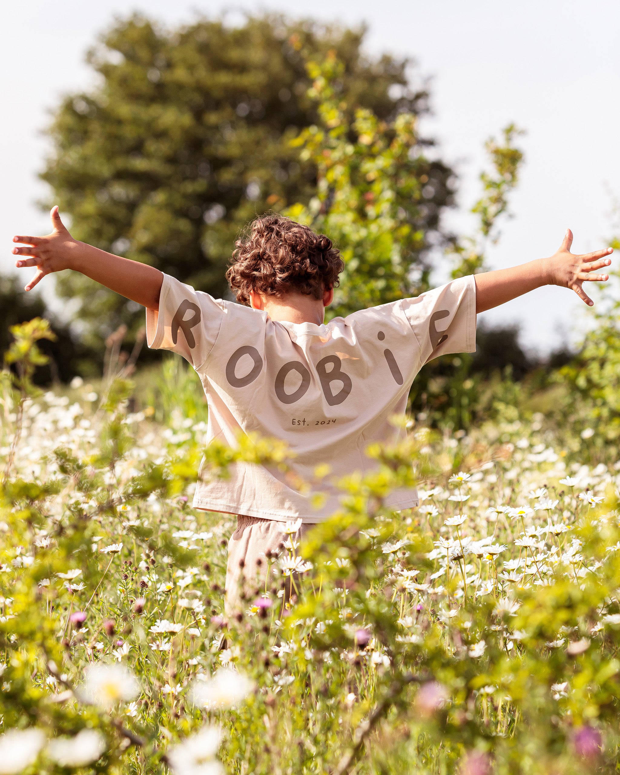 A child seen from behind in a sunlit field of white flowers, wearing a cream-colored oversize t-shirt with 'ROOBIE EST. 2024' printed in dark brown across the back.