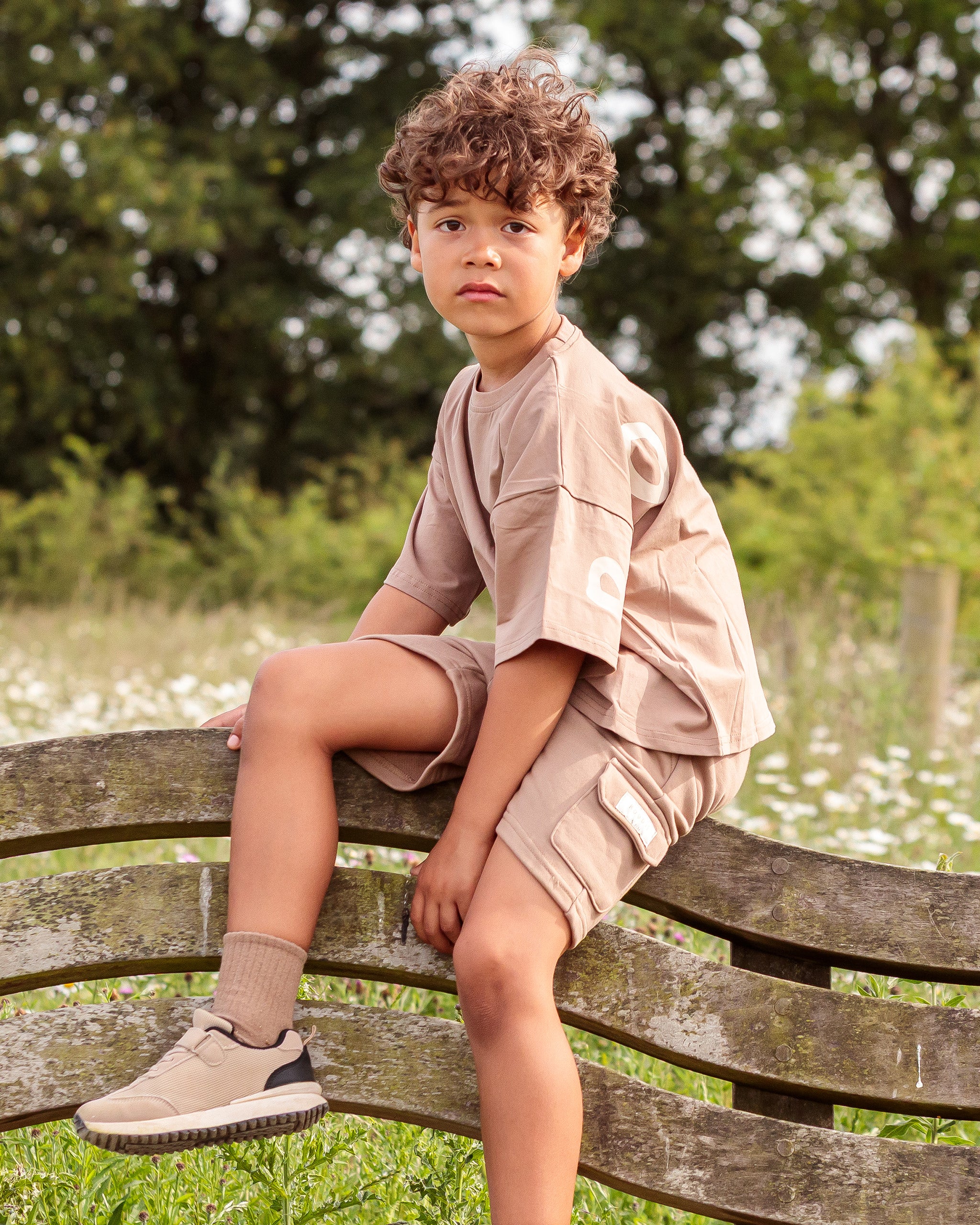 A young boy with curly brown hair sits on a wooden park bench wearing a matching brown co-ord with oversized t-shirt and cargo shorts set, paired with beige socks and sneakers, against a blurred green background.