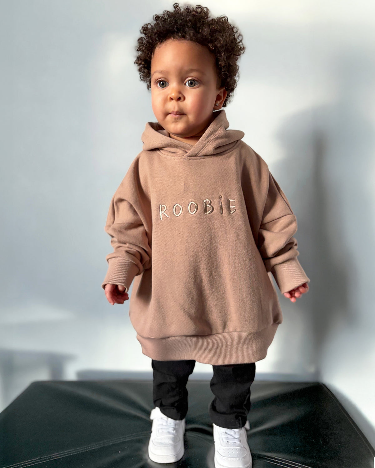 Child wearing a brown oversized hoodie with 'ROOBiE' printed on it, standing on a black surface with a white background.