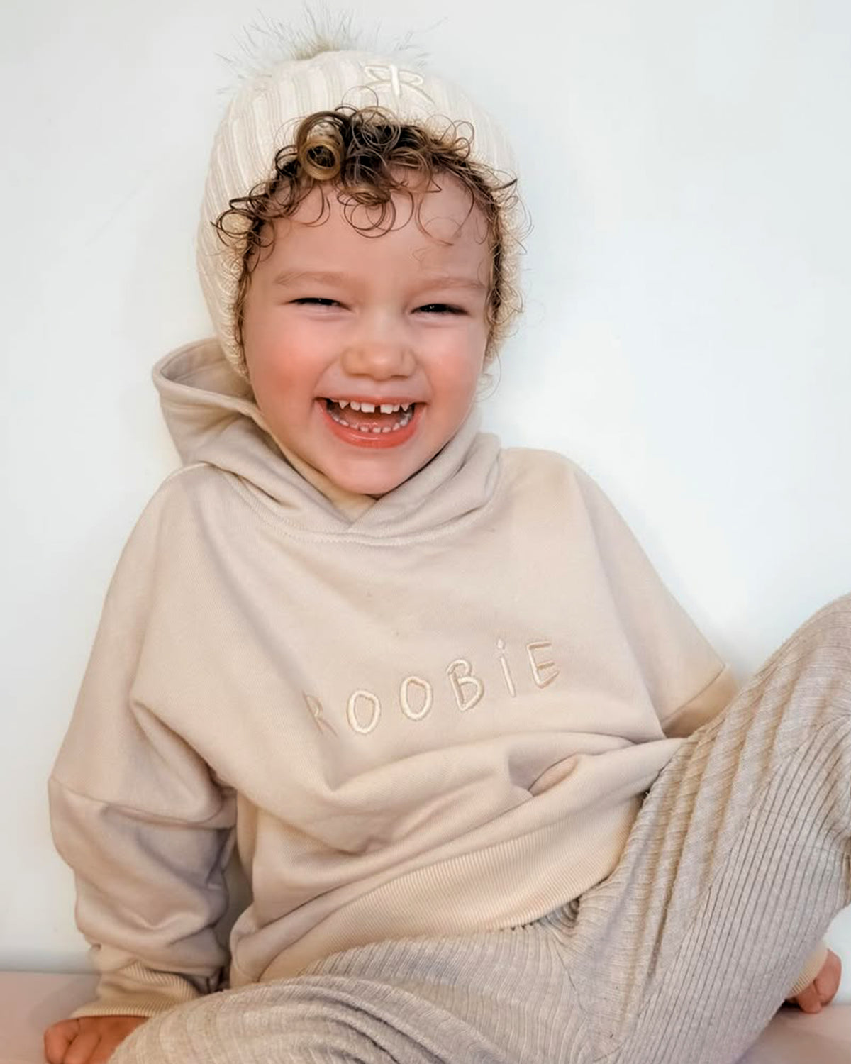 Child wearing a beige 'ROOBiE' oversized hoodie, ribbed neutral leggings and a matching knit bobble beanie on a white background