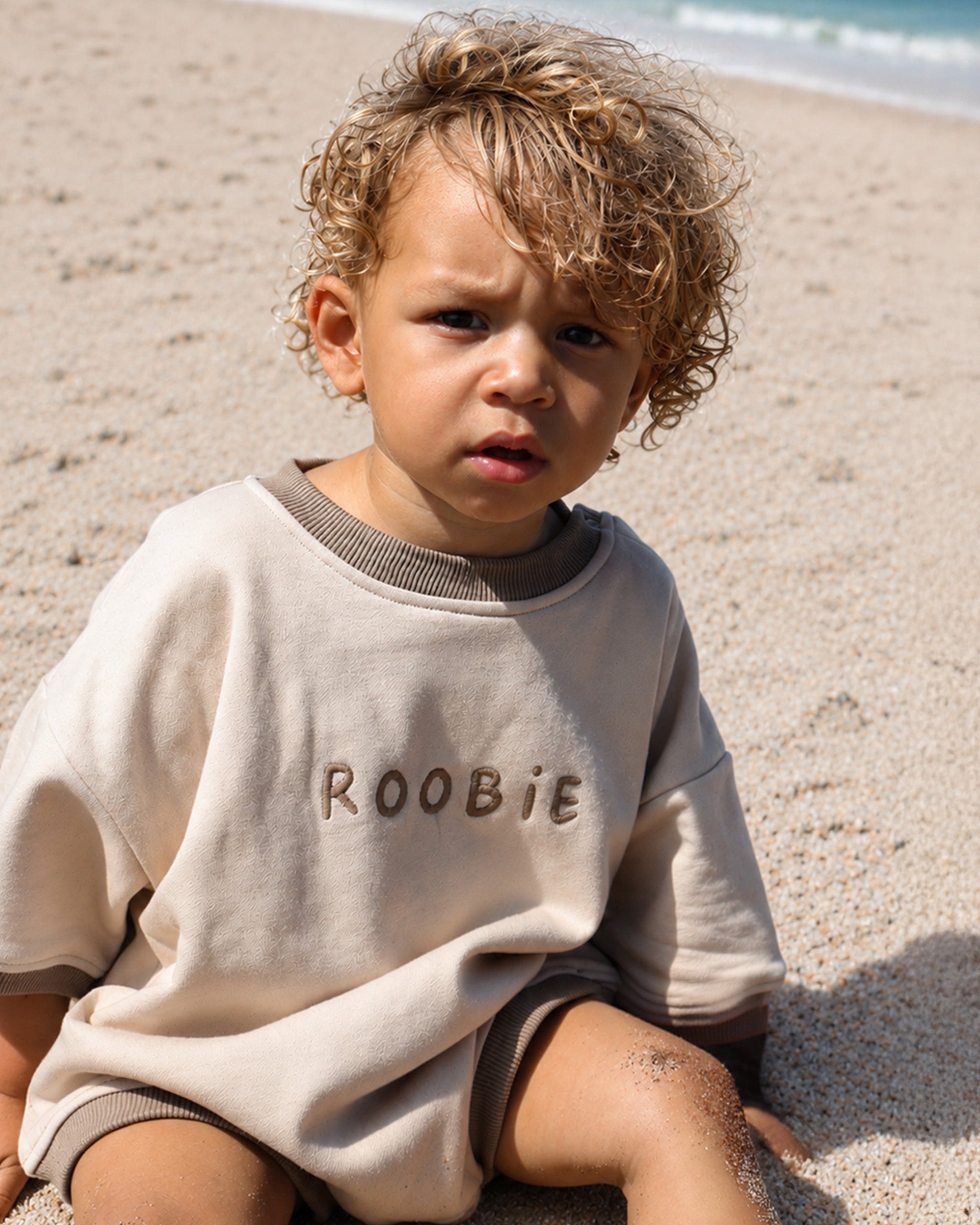 A toddler with curly blond hair sits on a sandy beach wearing an oversized cream Romper with the name 'ROOBiE' embroidered on the front.