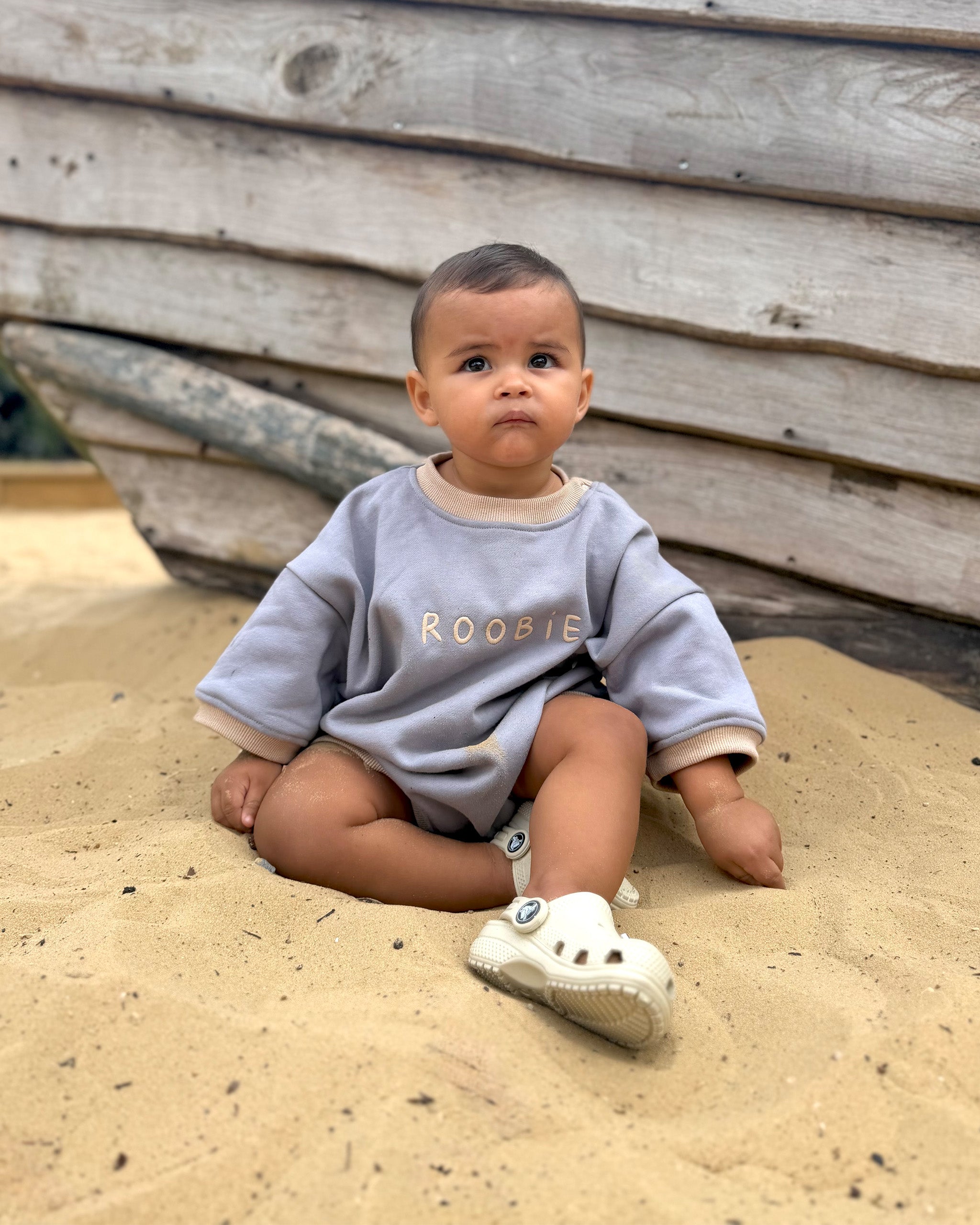 A baby sitting in sand wearing a light gray-blue Oversized Romper with 'ROOBIE' printed in cream across the chest, paired with white Crocs.