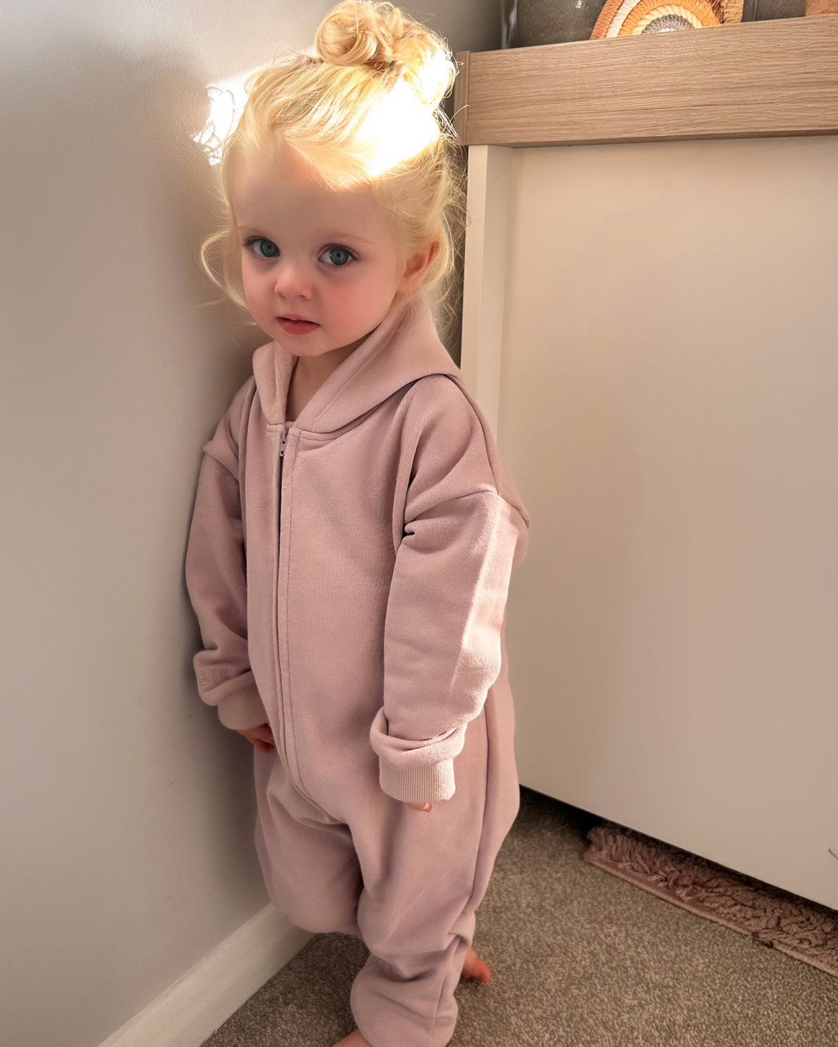 Child wearing a pink hooded one-piece standing in a room with neutral walls.