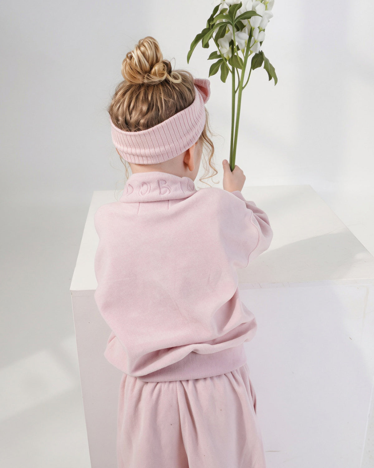 A child seen from behind wearing a light pink ribbed bow headband and a matching pale pink quarter zip jumper and wide leg jogger set, holding a green stem with white flowers against a white studio background.