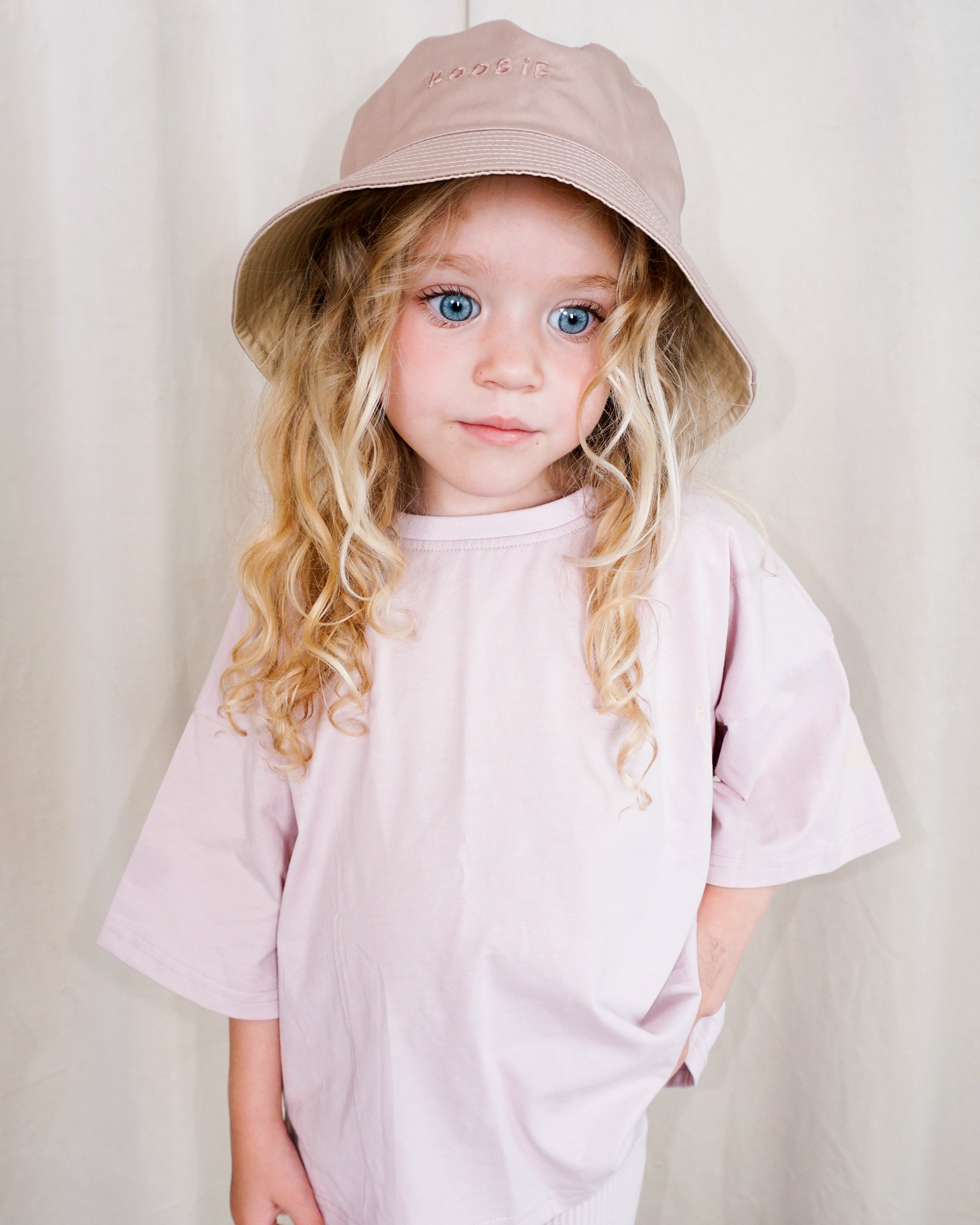 A young girl with long blonde curly hair wearing a dusty pink and cream reversible bucket hat and a matching light pink oversized t-shirt, standing against a neutral cream-colored curtain backdrop.