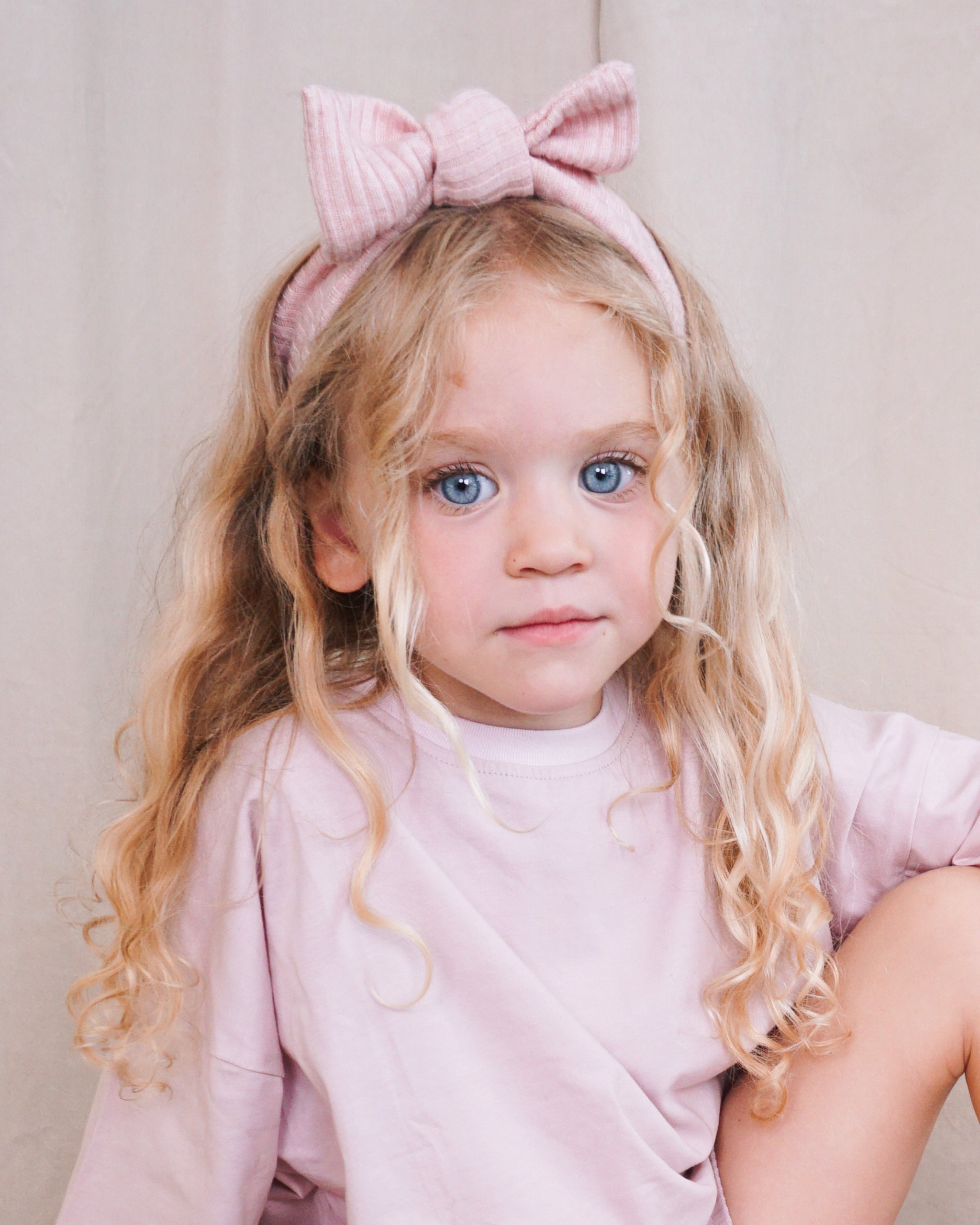 A young girl with long blonde curly hair wearing a light pink ribbed fabric headband with a large bow, paired with a matching pale pink oversized t-shirt, posed against a neutral beige background.