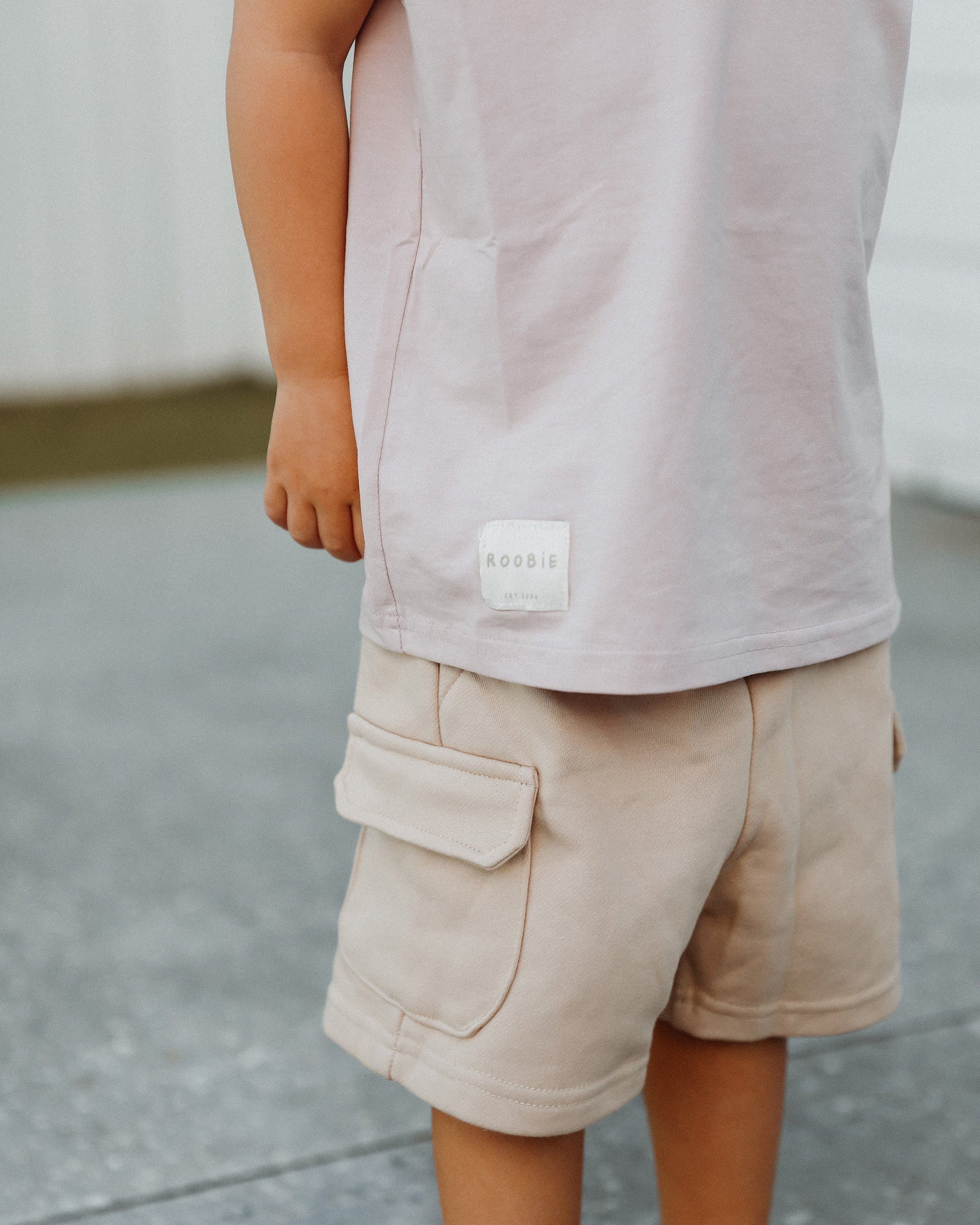 A child wearing a light pink Singlet t-shirt and beige cargo shorts, shown from the waist down. The shirt has a small white square patch on the lower left side with the brand name 'ROOBiE' printed on it.