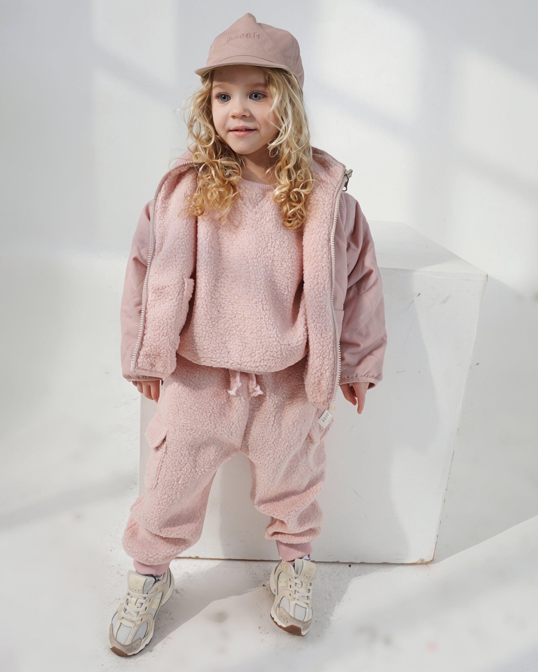 Toddler with pink curly hair wearing a reversible cream and pink baseball cap with matching pink teddy jumper and cargos with a reversible 4-in-1 coat with a white studio backdrop.