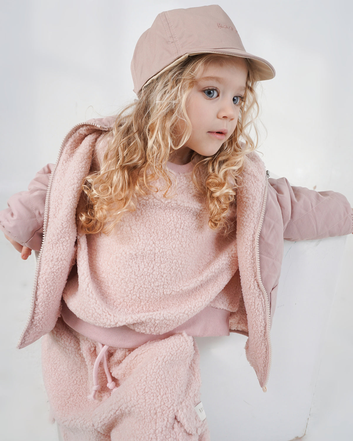 A young girl with long curly blonde hair wearing a dusty pink teddy tracksuit, matching reversible cap, and 4-in-1 reversible coat, posing against a white wall.