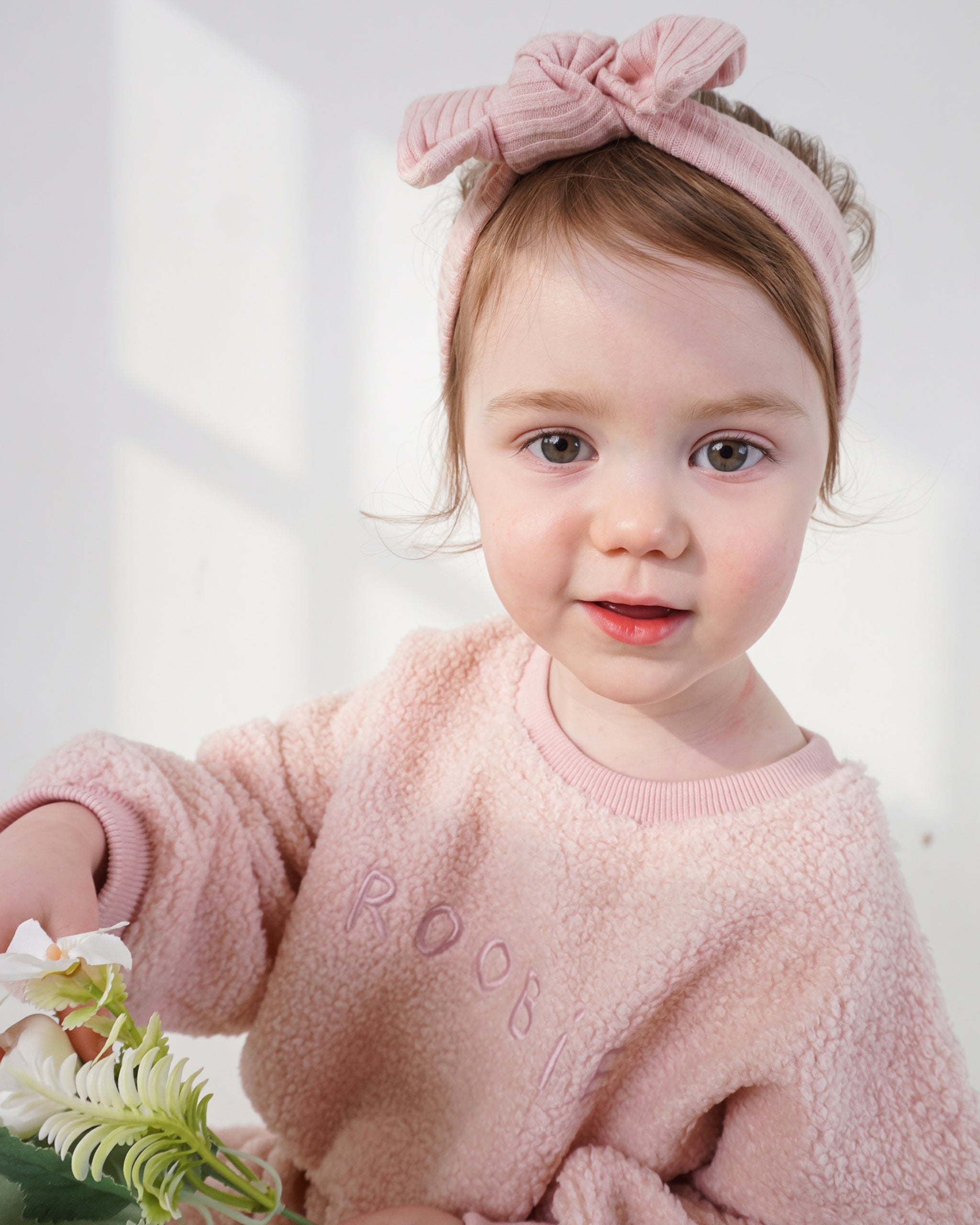 A toddler girl wearing a dusty pink teddy one-piece and a matching pink ribbed bow headband, holding white flowers against a bright white background.