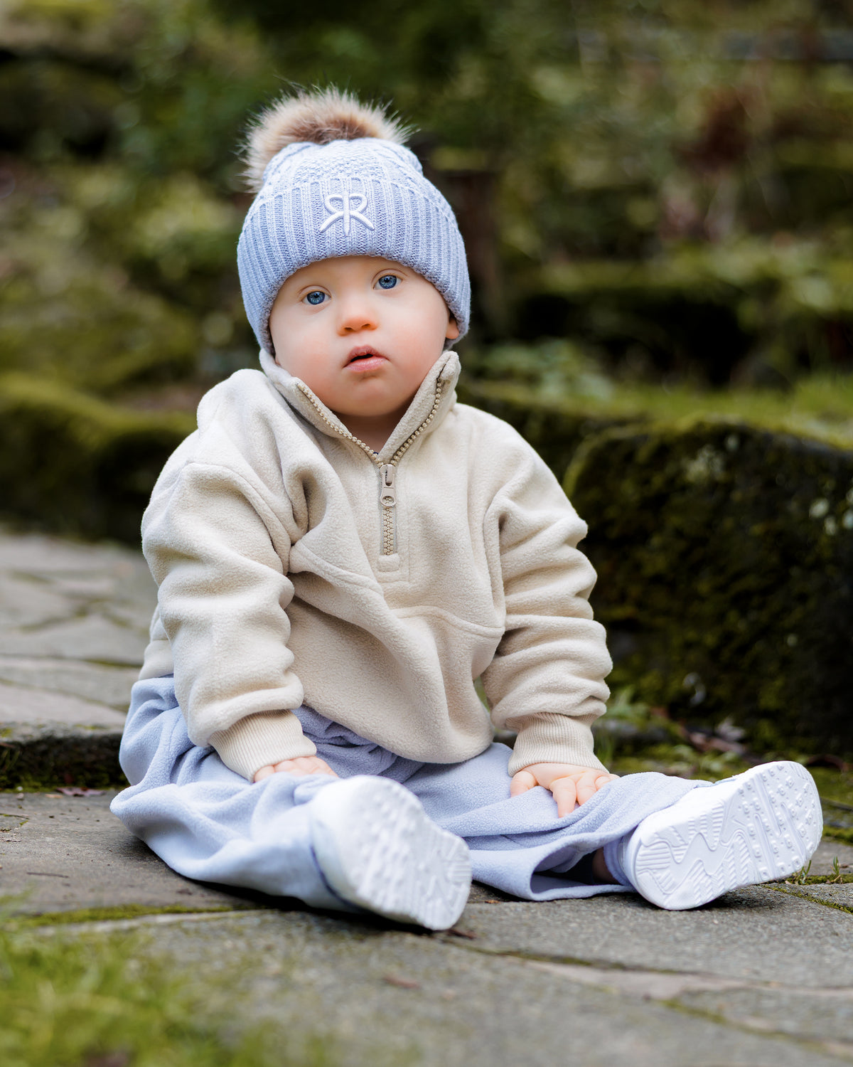 Baby sitting outdoors wearing a beige quarter zip fleece jumper with light blue wide leg joggers and matching knit bobble beanie with a pom-pom.