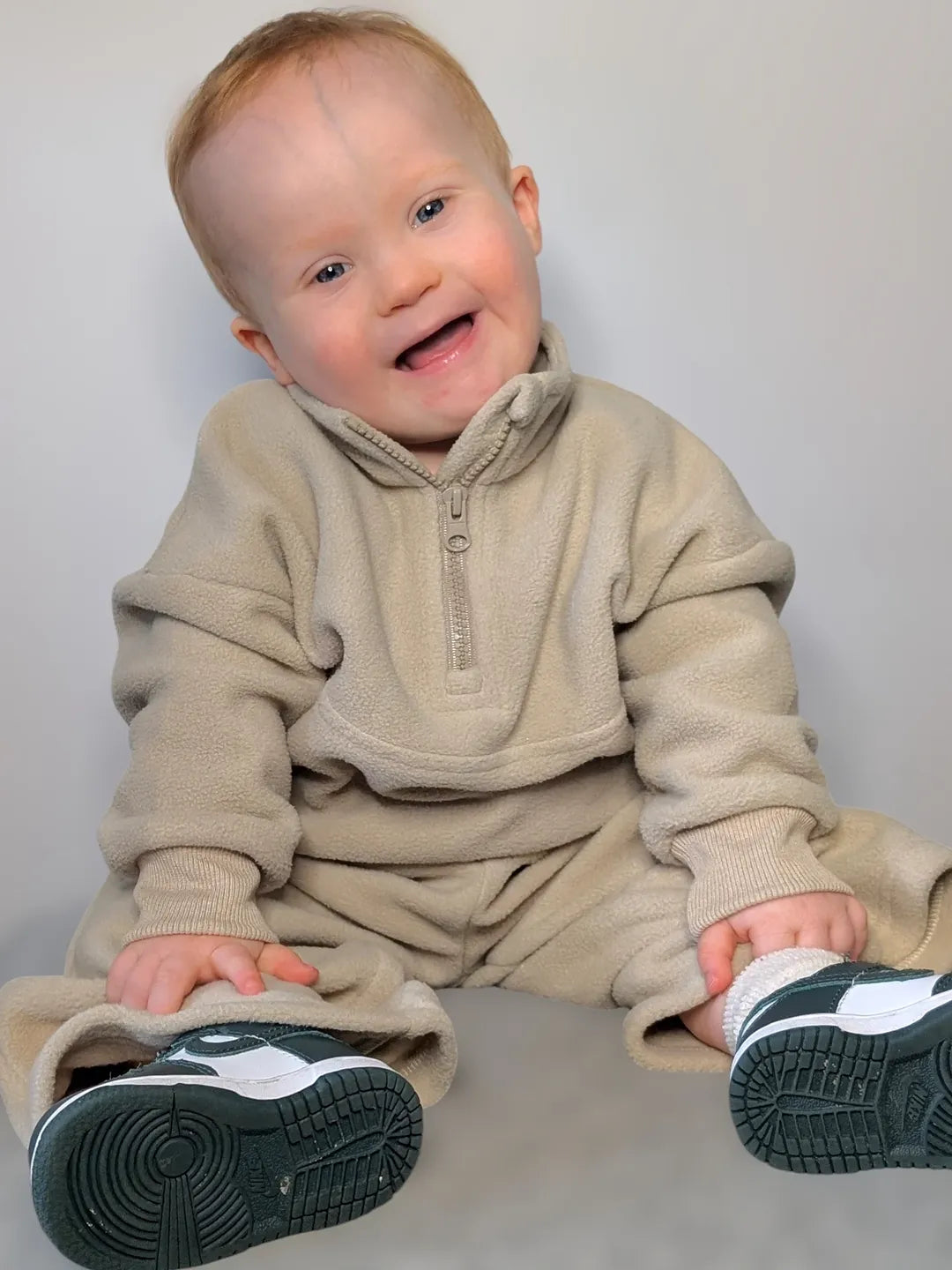 Baby wearing a beige fleece quarter zip with matching wide leg joggers outfit with black and white shoes on a plain background