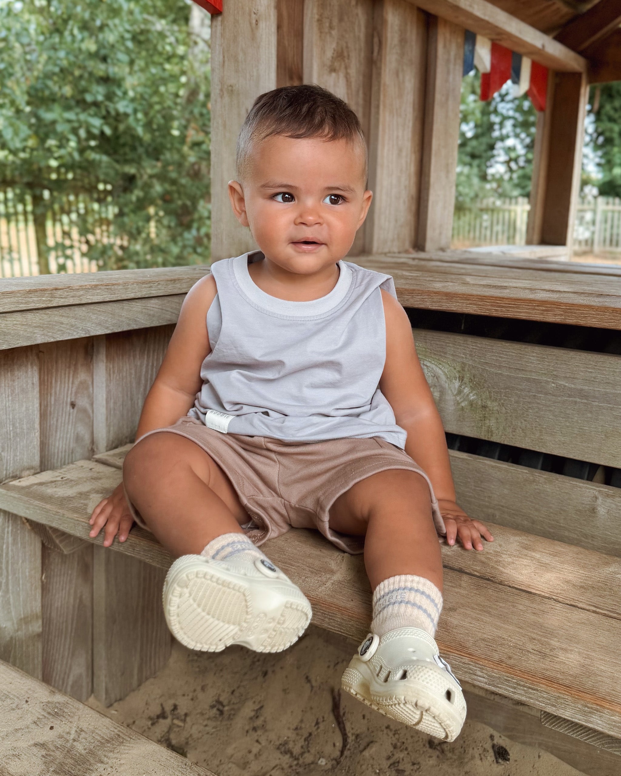 A toddler boy sitting on a wooden bench at an outdoor pavilion, wearing a light gray/blue (BONDi BREEZE) sleeveless Singlet top, tan (CHOCCY BiCCY) Cargo Shorts, ROOBiE cream and blue crew striped socks, and white croc style shoes.
