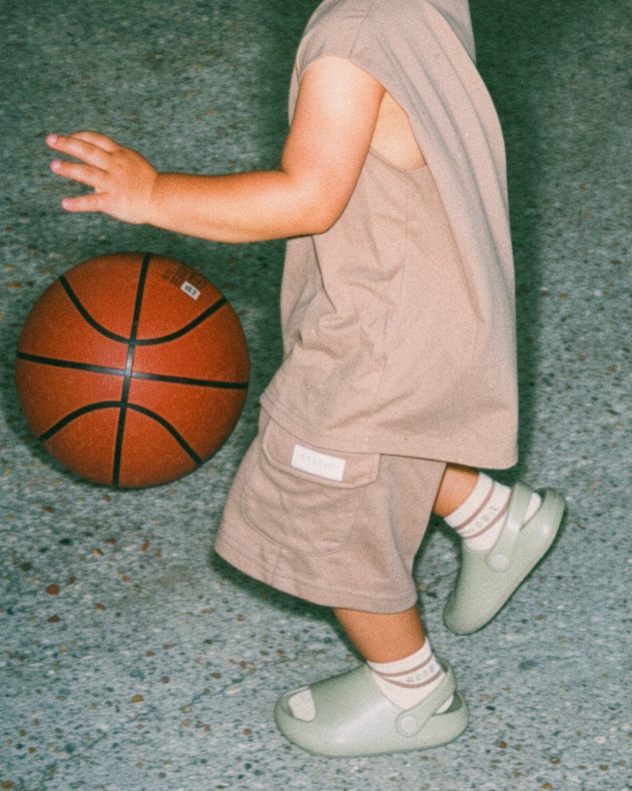 A child wearing a brown (CHOCCY BiCCY) sleeveless Singlet top and Cargo Shorts with light green slip-on shoes and ROOBiE Socks, standing next to a basketball on a speckled gray floor.