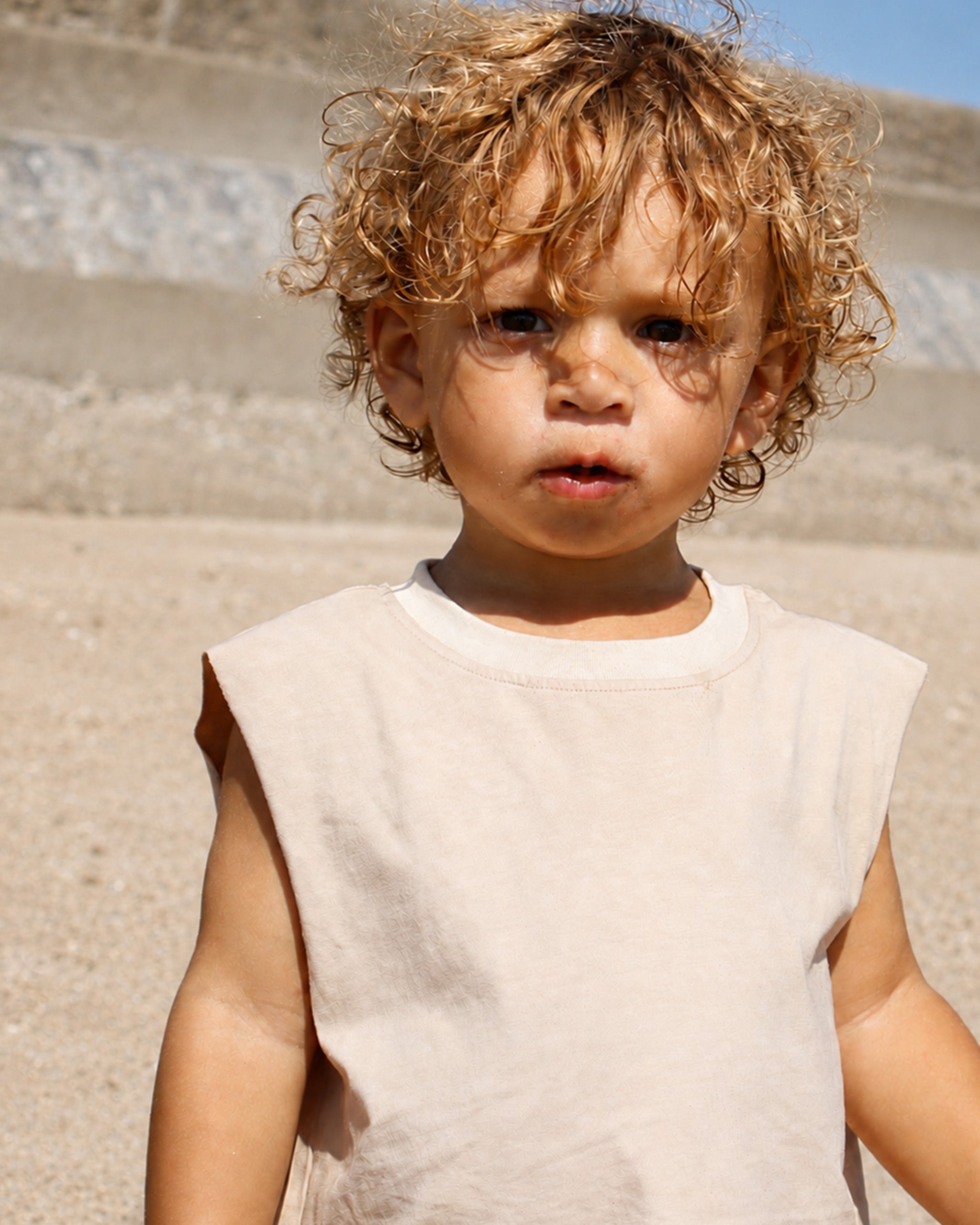 A young child with curly light brown hair wearing a sleeveless oversized beige Singlet t-shirt, standing outdoors against a neutral, sandy, sunlit background.