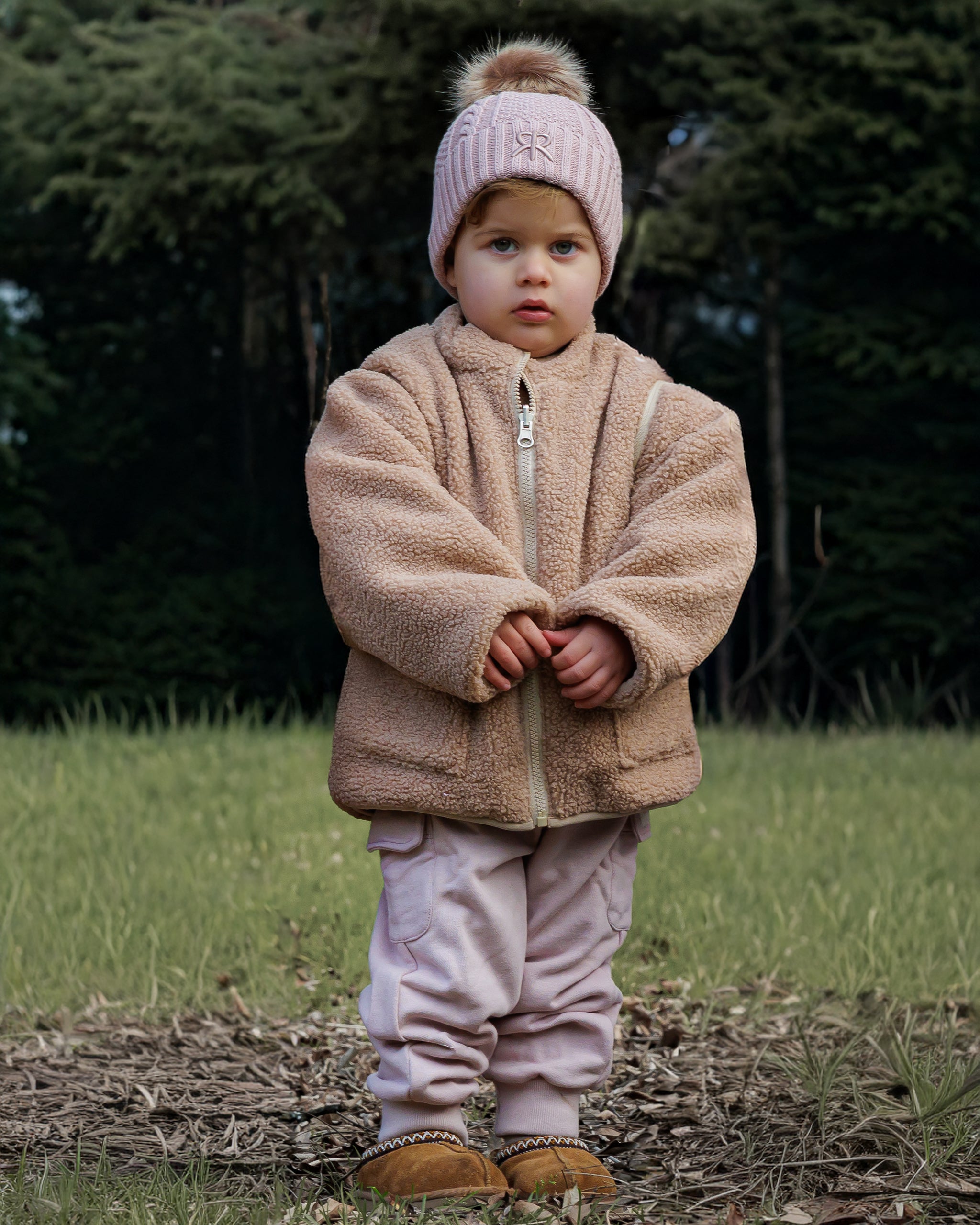 Child wearing a brown 4-in-1 reversible teddy fleece jacket and pink knit bobble bobble with matching pink cargos outdoors