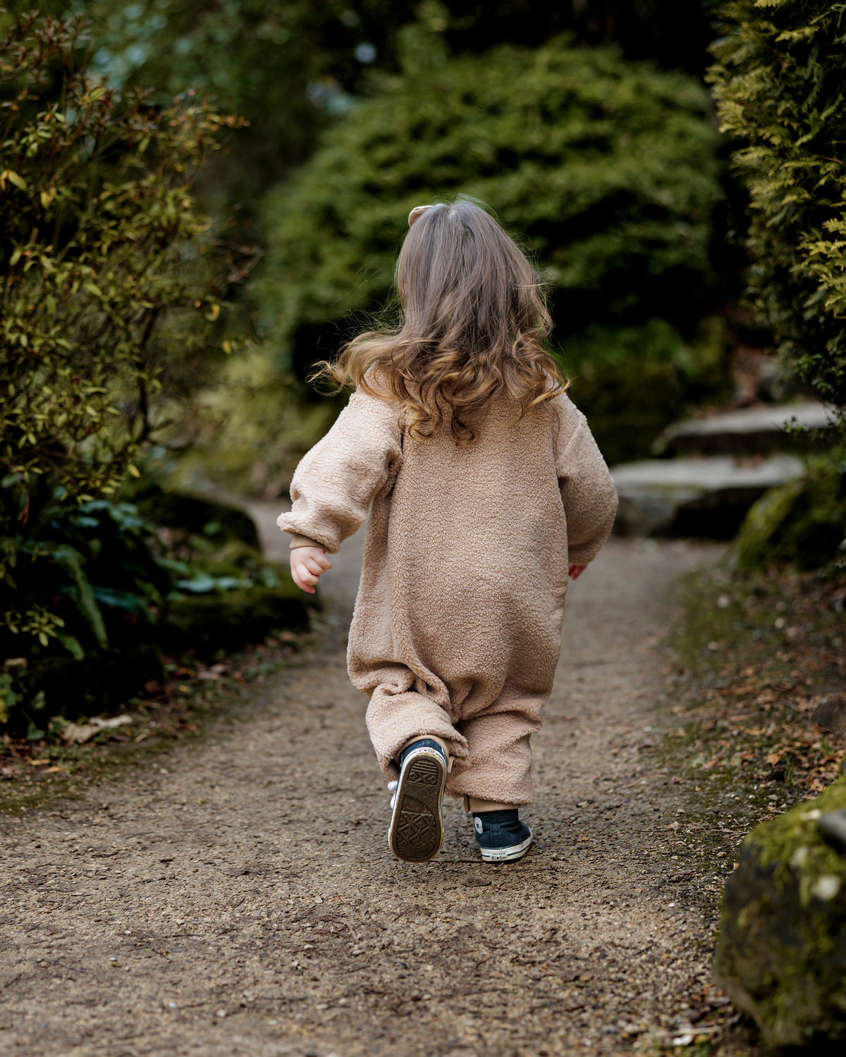 Child in a brown teddy onesie walking away on a path through a forest