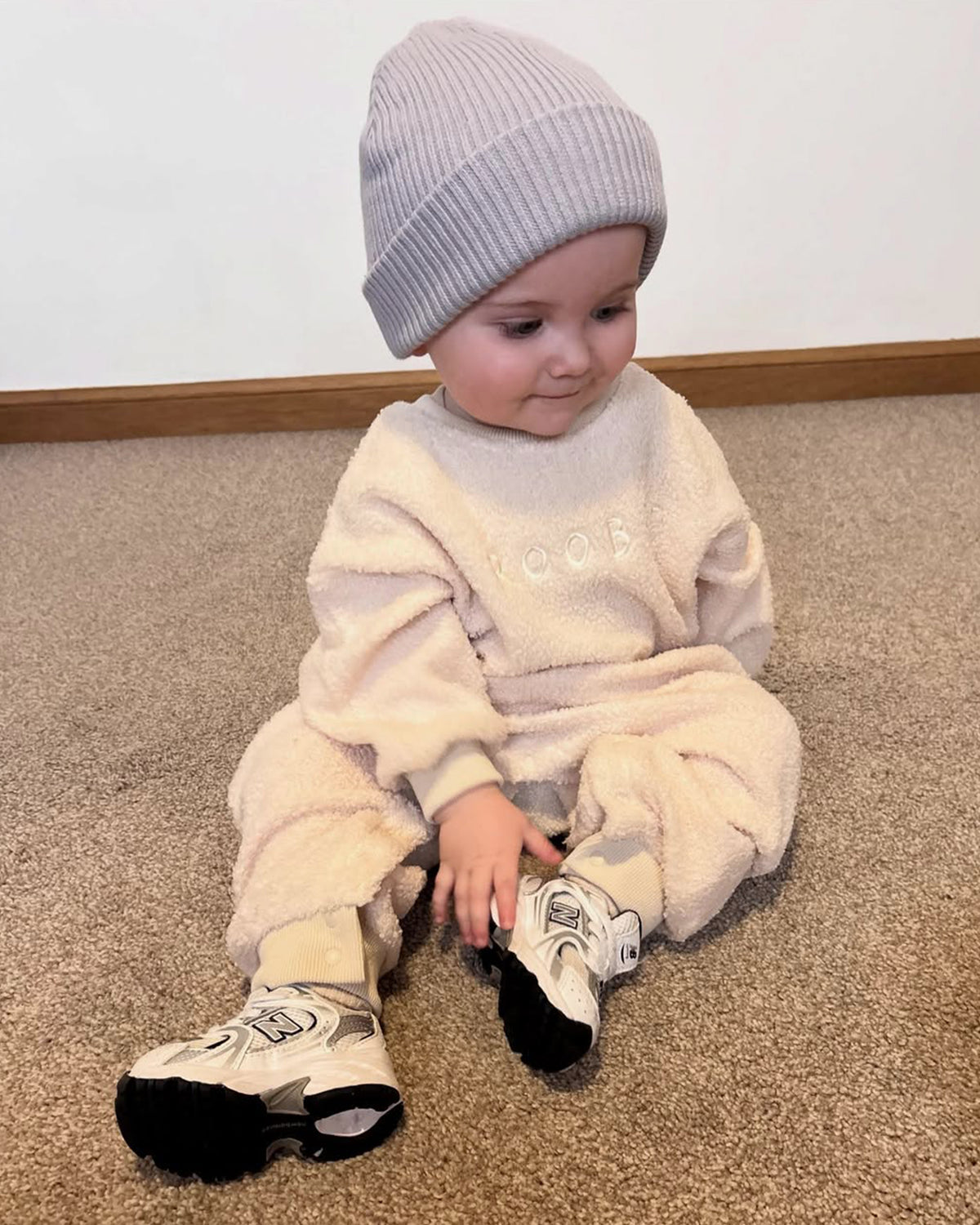 Child wearing a cream teddy one-piece outfit with a blue cotton ribbed beanie hat, sitting on a carpeted floor.