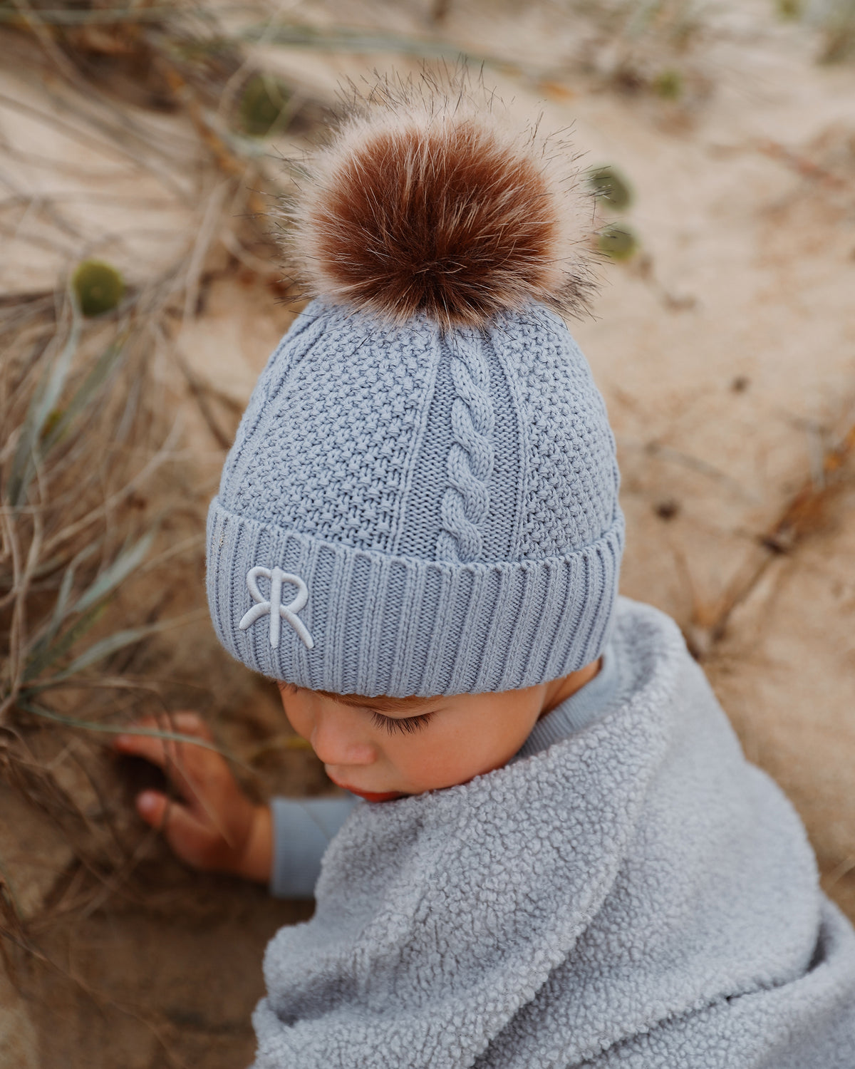 A baby wearing a light blue cable-knit bobble beanie with a brown faux fur pom-pom and embroidered logo detail, wearing in a matching teddy fleece jumper in a sandy outdoor setting.
