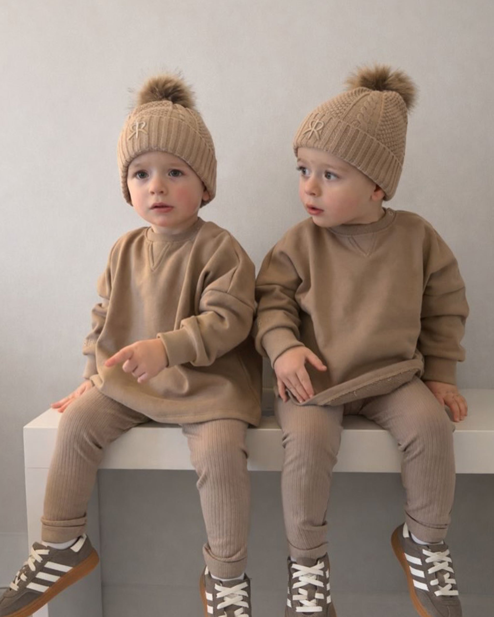 Twin boys wearing matching brown winter outfits with jumper, ribbed leggings and knit bobble beanie and shoes, sitting on a white surface.