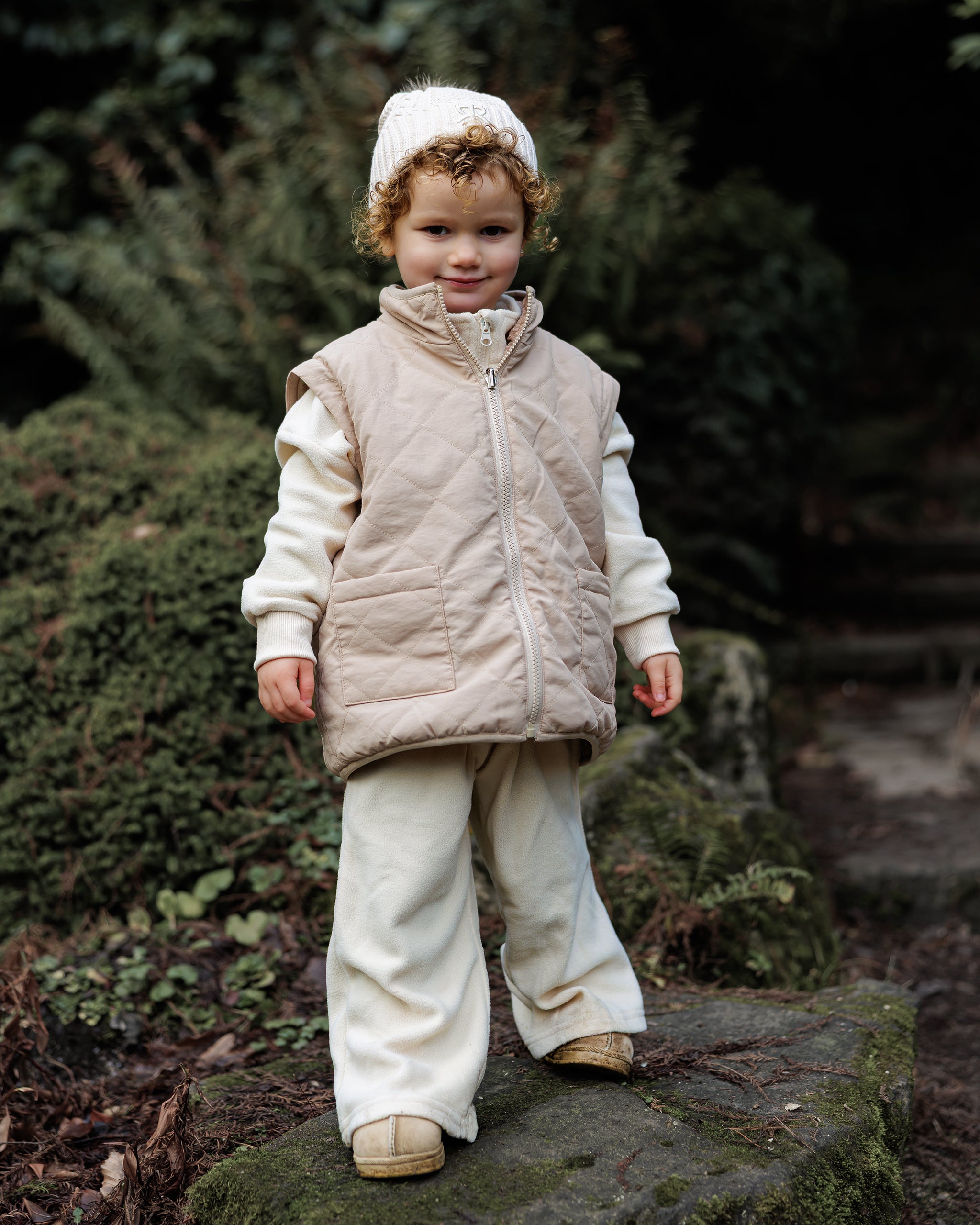 Child wearing a beige quilted gilet from 4-in-1 reversible teddy coat and cream wide leg joggers standing outdoors with a cream knit bobble beanie with pom pom.