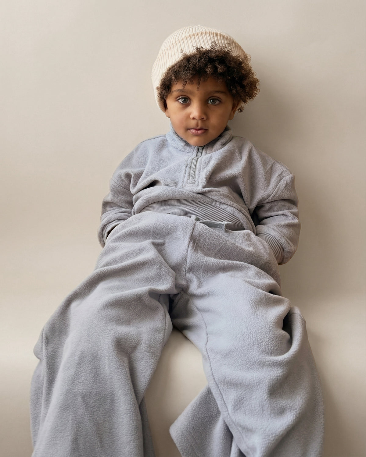 Child wearing a light blue tracksuit and beige beanie sitting on a neutral surface.