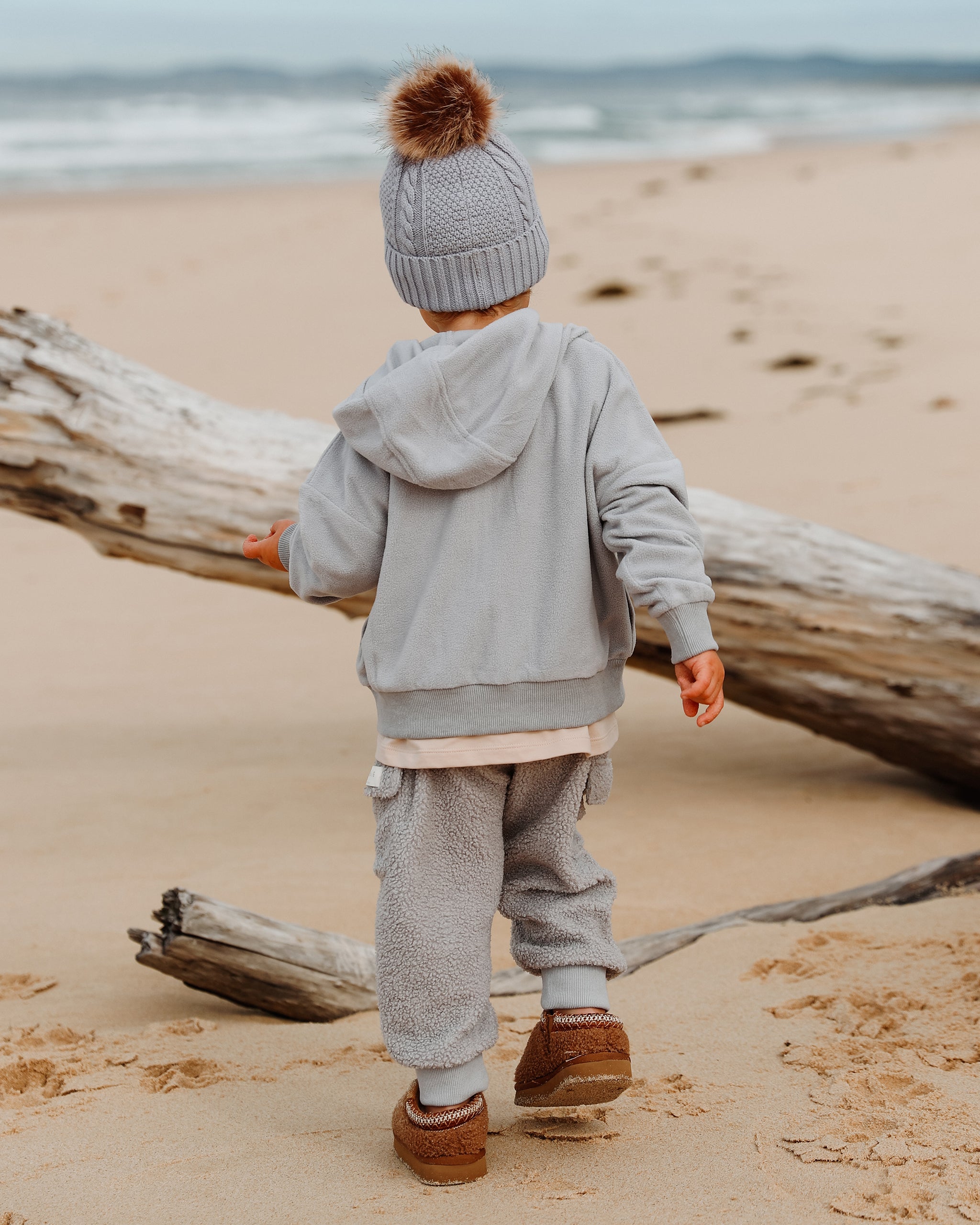 A toddler seen from behind on a sandy beach, wearing a light gray-blue knit bobble beanie with a brown pom-pom, a matching fleece zip up hooded sweatshirt and blue-gray neutral wide jogger pants co-ord, and brown slip-on shoes, with driftwood and the ocean in the background.