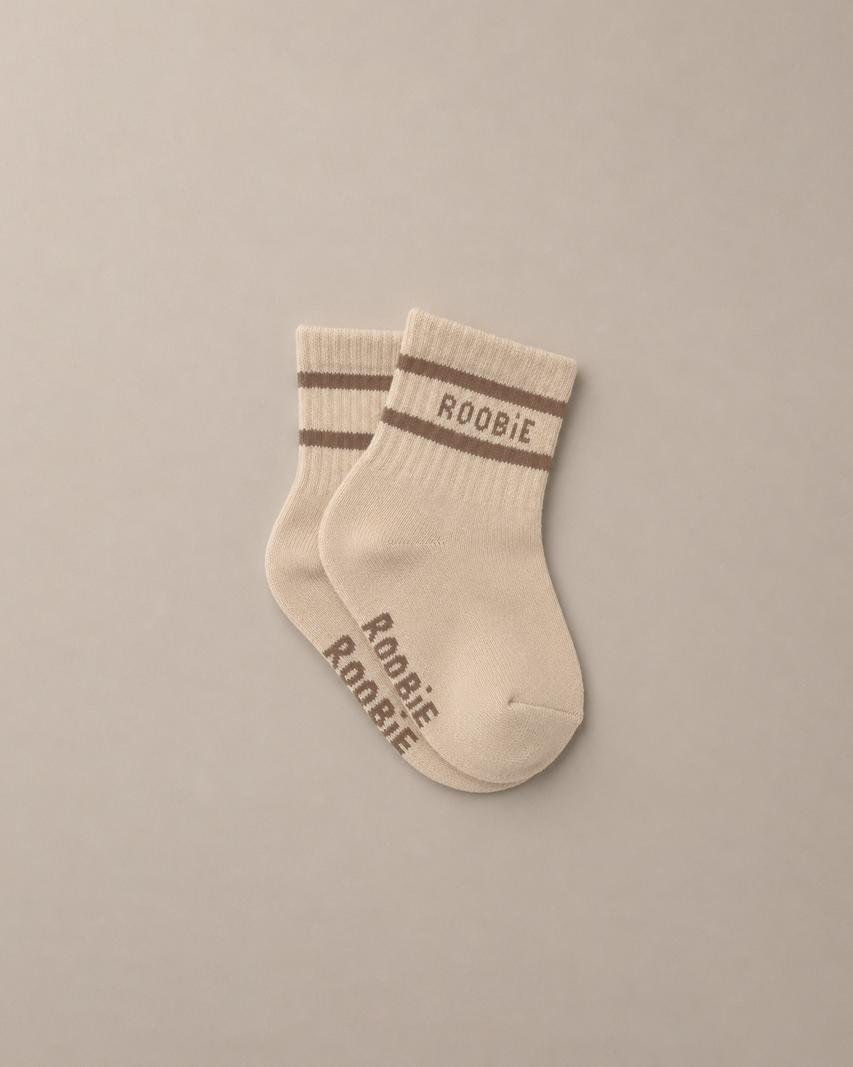 A pair of cream-colored baby socks with brown stripes and 'ROOBIE' printed in brown on the cuff and foot, displayed on a neutral beige background.