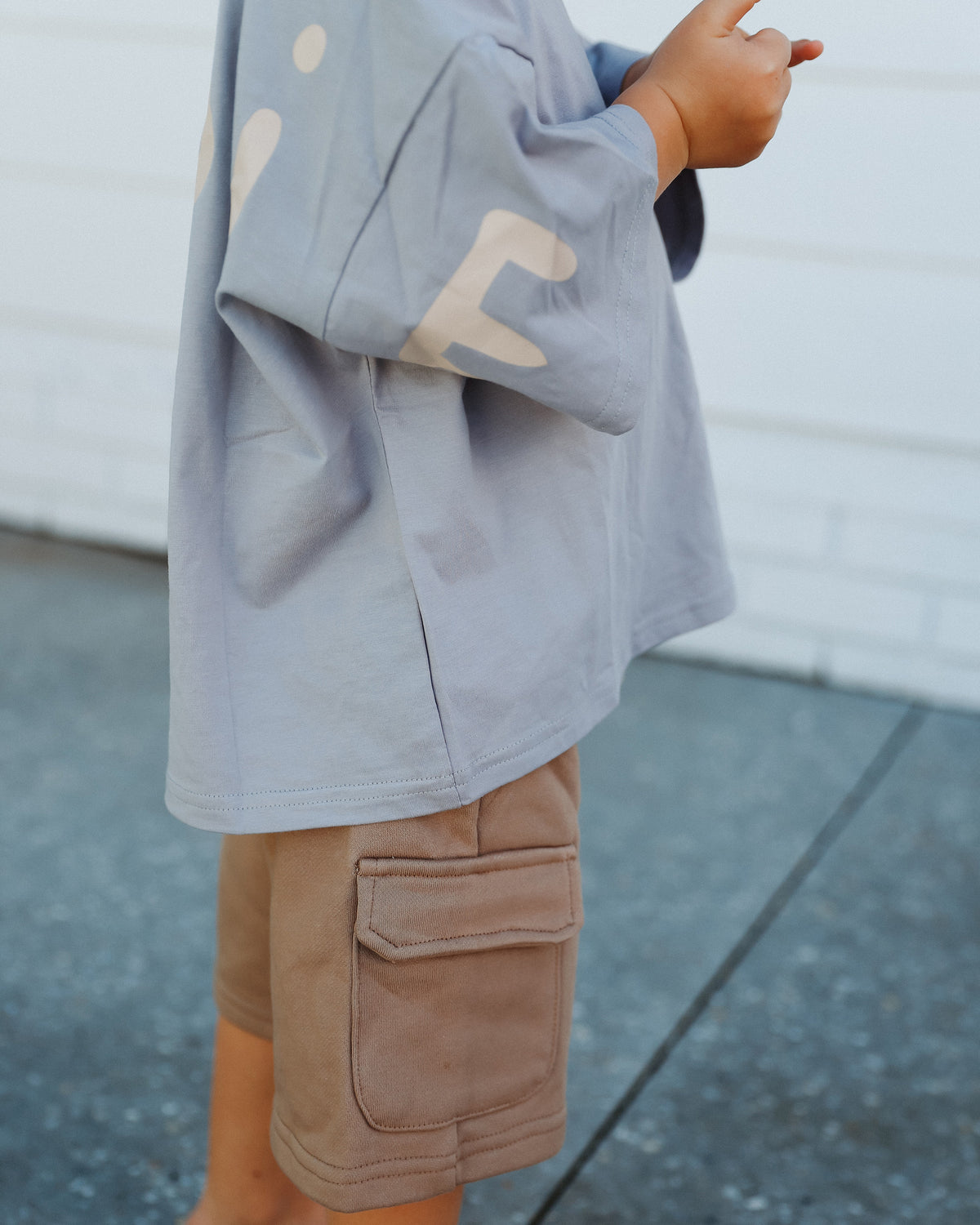 A child wearing a light blue oversized t-shirt with beige 'ROOBiE' lettering and tan neutral cargo shorts, standing outdoors against a white wall at nursery.