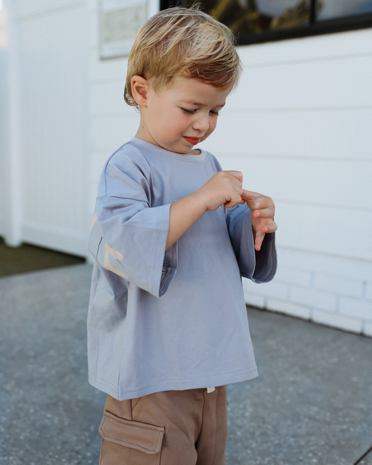 A young child mixing and matching a light blue Oversized T-shirt and brown Cargo Shorts, standing outdoors in front of a neutral white building.