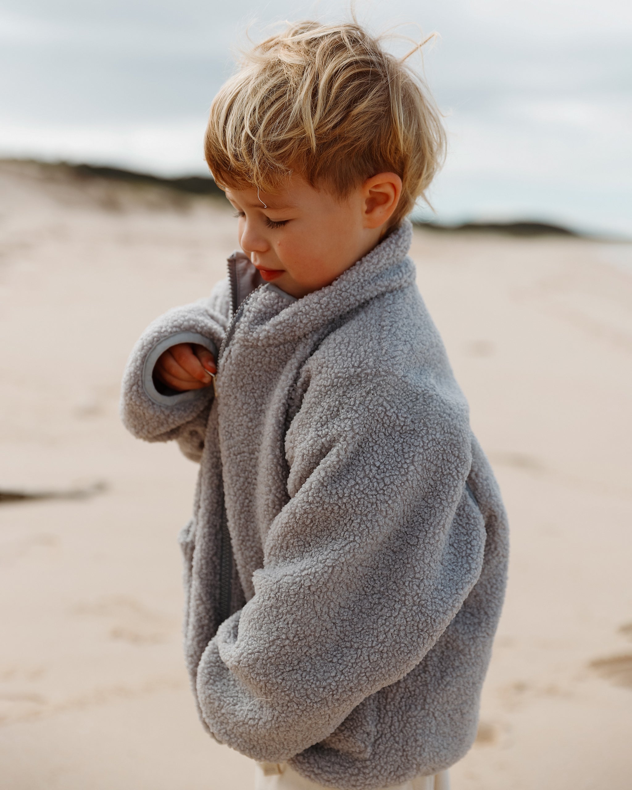 A young boy with blonde hair stands on a sandy beach wearing a light neutral blue-gray teddy fleece 4-in-1 reversible coat.