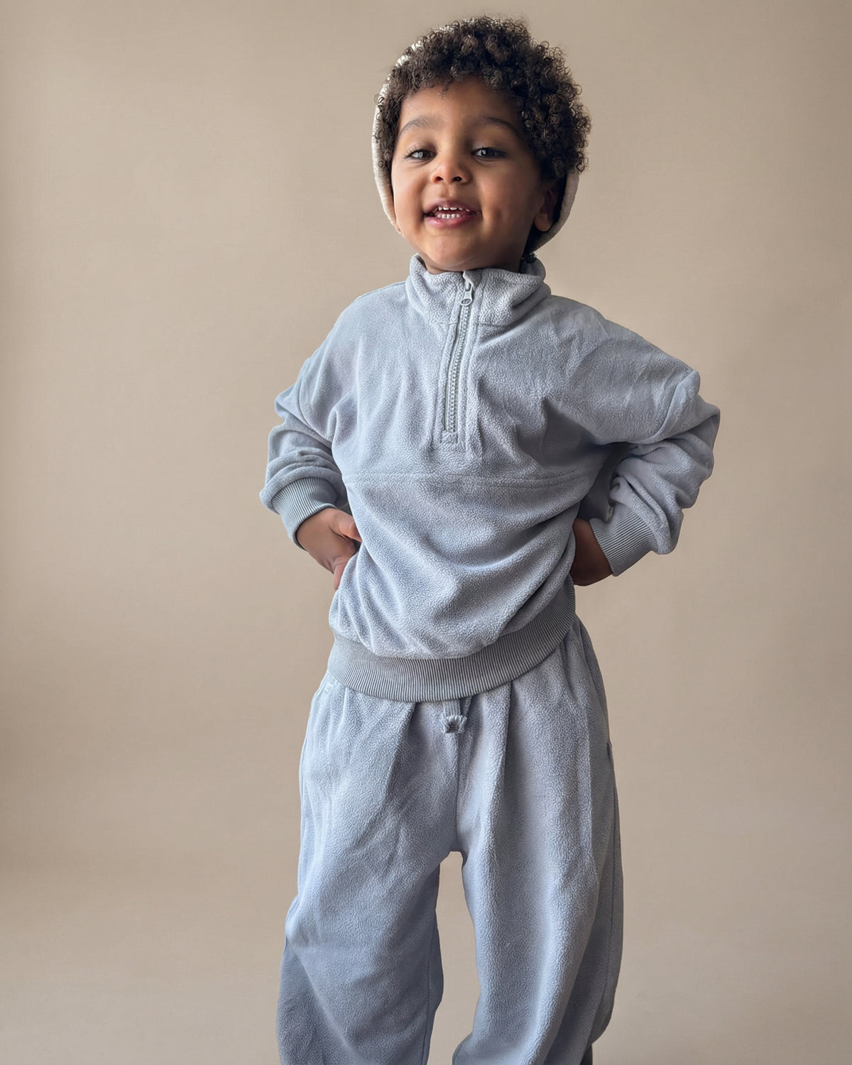 Child wearing a light blue quarter zip tracksuit with wide leg joggers with cream cotton ribbed beanie against a beige background